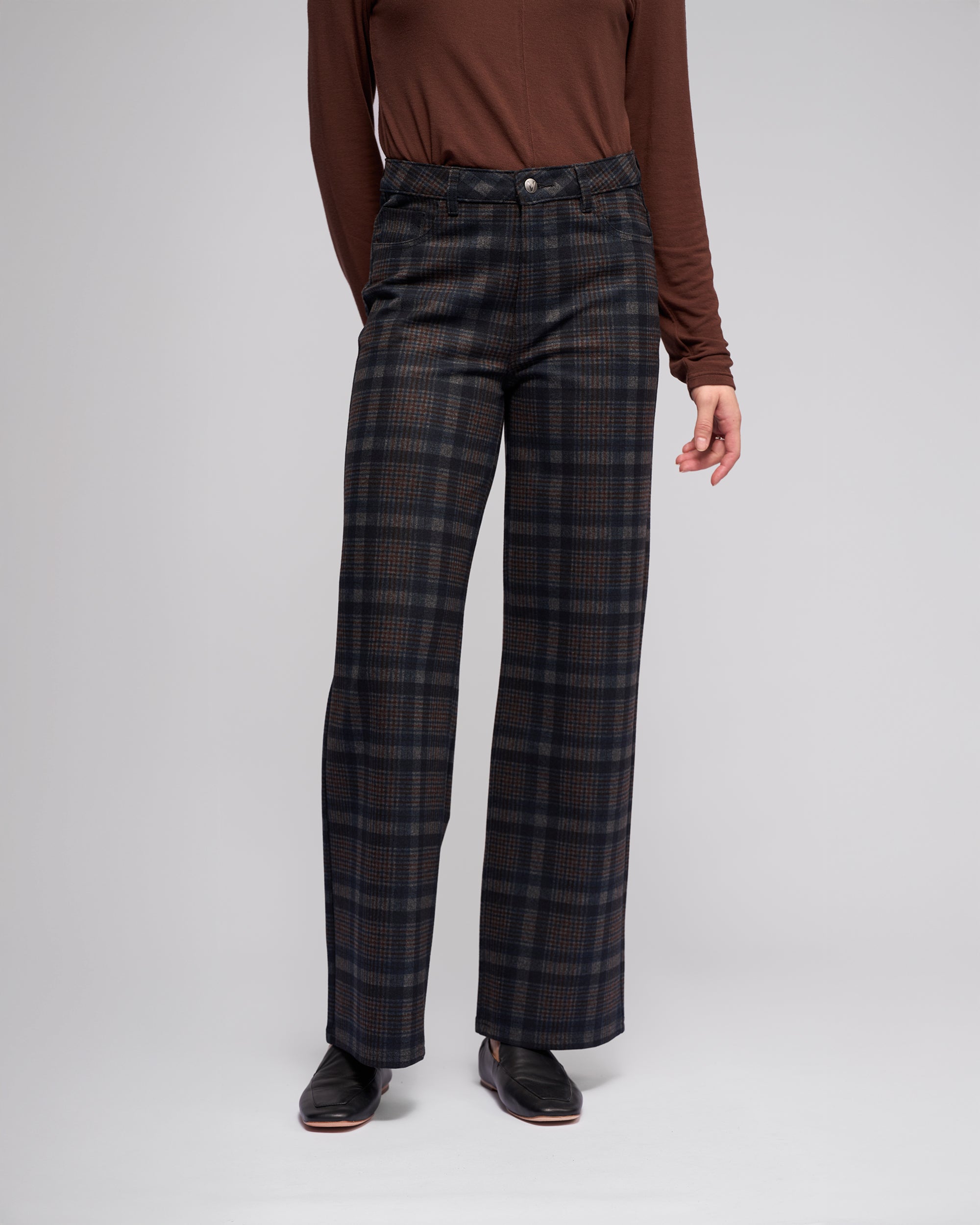 A person stands against a plain light background wearing a long-sleeve brown shirt, Vassalli Empire Pant - Nomad in dark tones, and black shoes. The person's head is not visible in the image.
