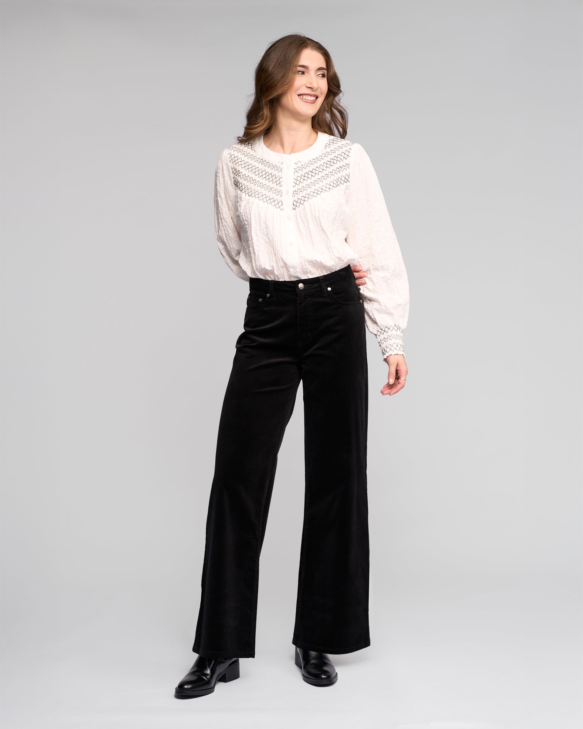 A woman smiles against a plain background, wearing a white lace-detailed blouse, black Vassalli Empire Cord Pant, and black ankle boots.
