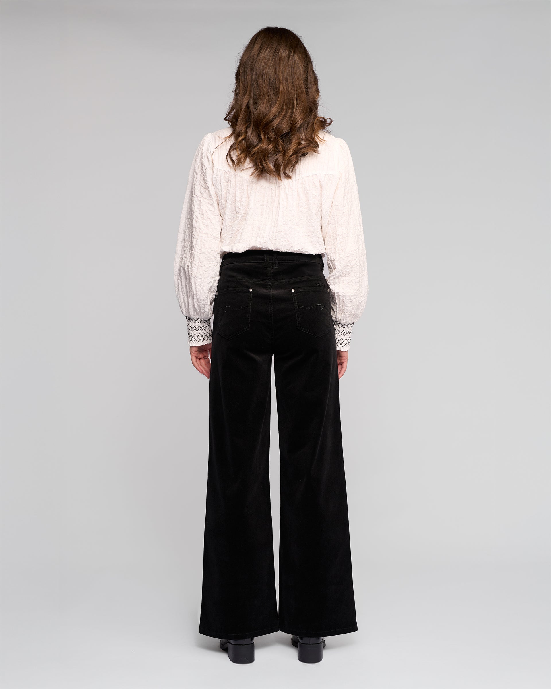 A woman with wavy brown hair, wearing a white blouse and Vassalli Empire Cord Pant in black, stands facing away from the camera against a plain light gray background.