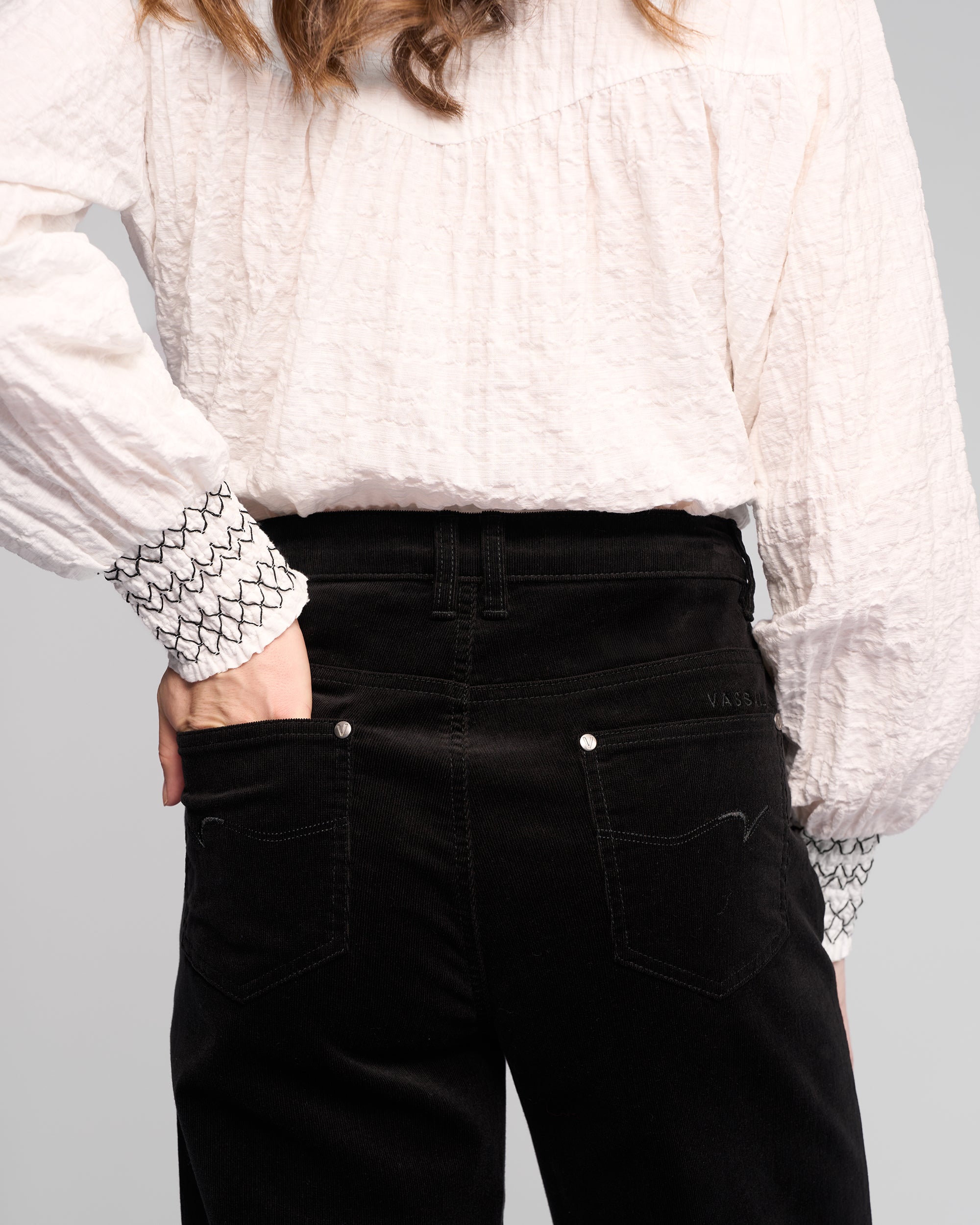 A person wears Vassalli’s Empire Cord Pant in black, styled high-waisted, paired with a textured white blouse with patterned cuffs, standing with one hand in a back pocket and facing away from the camera.