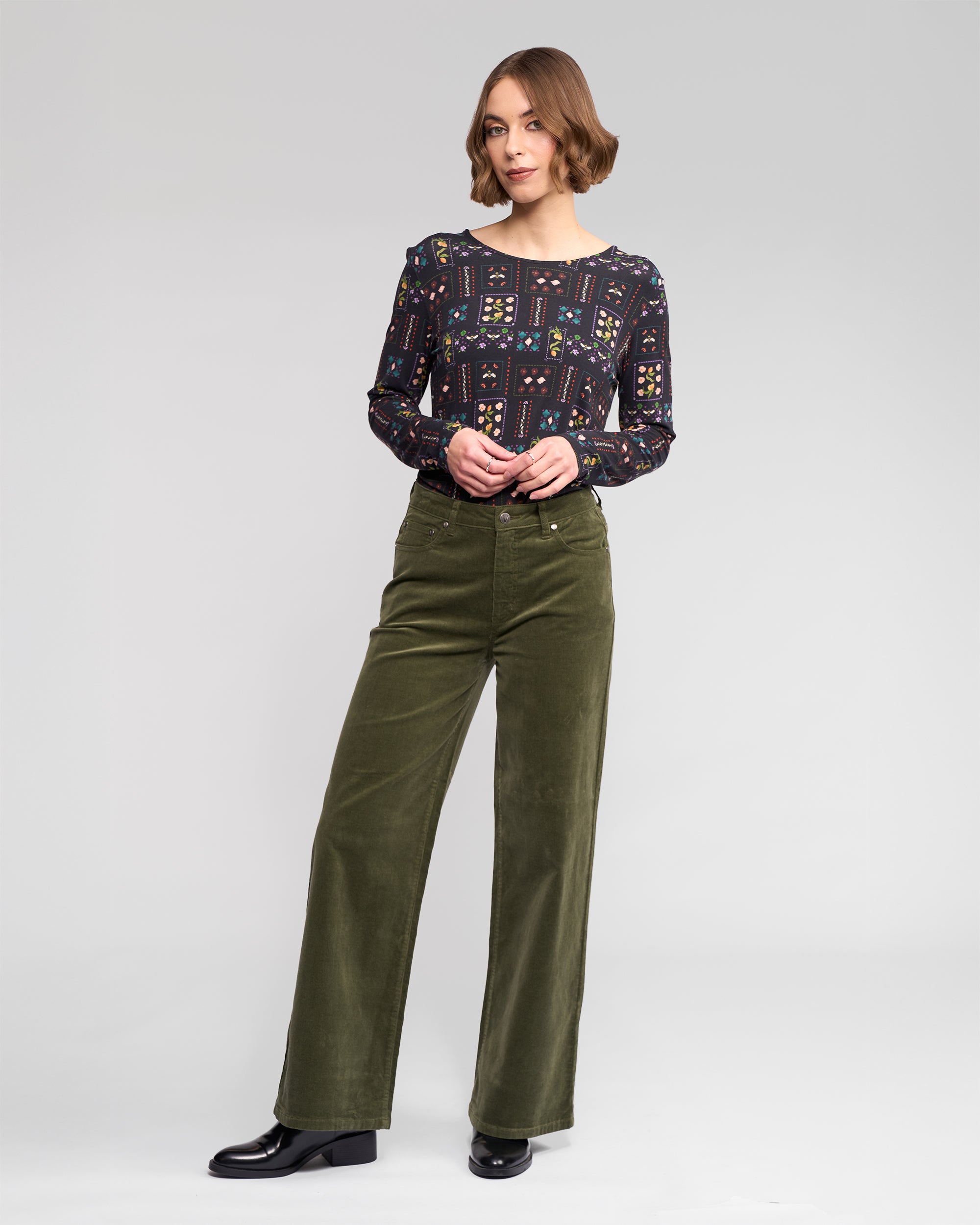A woman with short brown hair wears the Vassalli Empire Cord Pant in Deep Sage, paired with a patterned long-sleeve blouse and black shoes. She stands against a plain light gray backdrop, looking to the side with one hand in her pocket—winter ready.