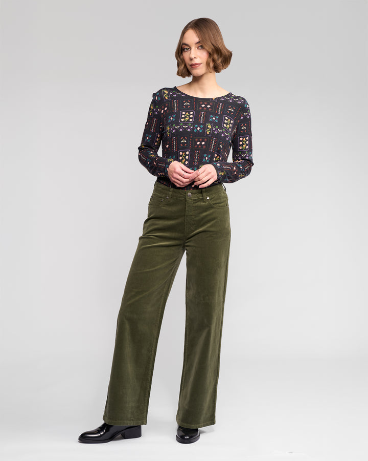 A woman with short brown hair wears the Vassalli Empire Cord Pant in Deep Sage, paired with a patterned long-sleeve blouse and black shoes. She stands against a plain light gray backdrop, looking to the side with one hand in her pocket—winter ready.