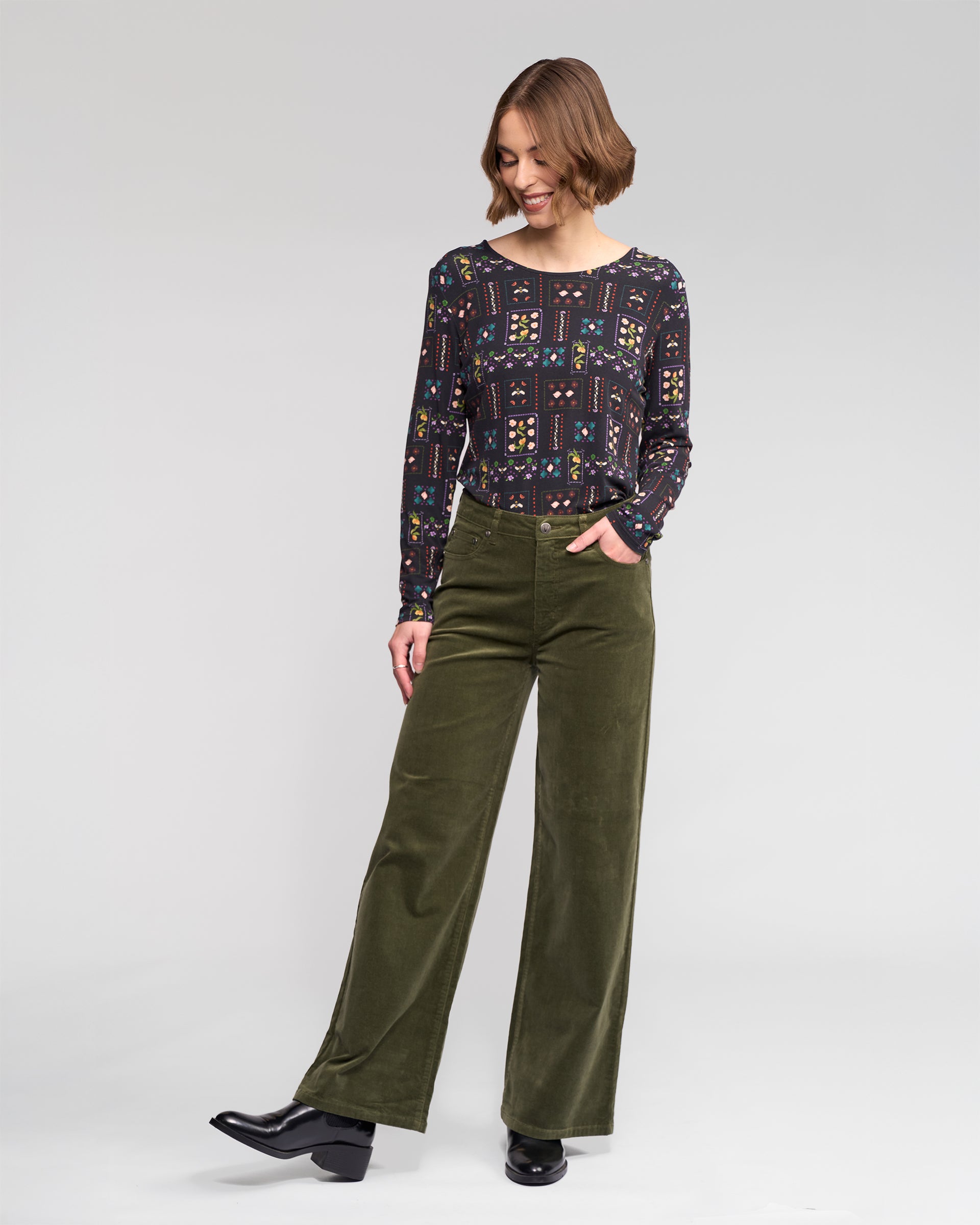 A woman with short brown hair smiles and looks to the side, wearing Vassalli's Empire Cord Pant in Deep Sage, a patterned long-sleeve top, and black heeled boots. She stands against a plain light background with one hand in her pocket.