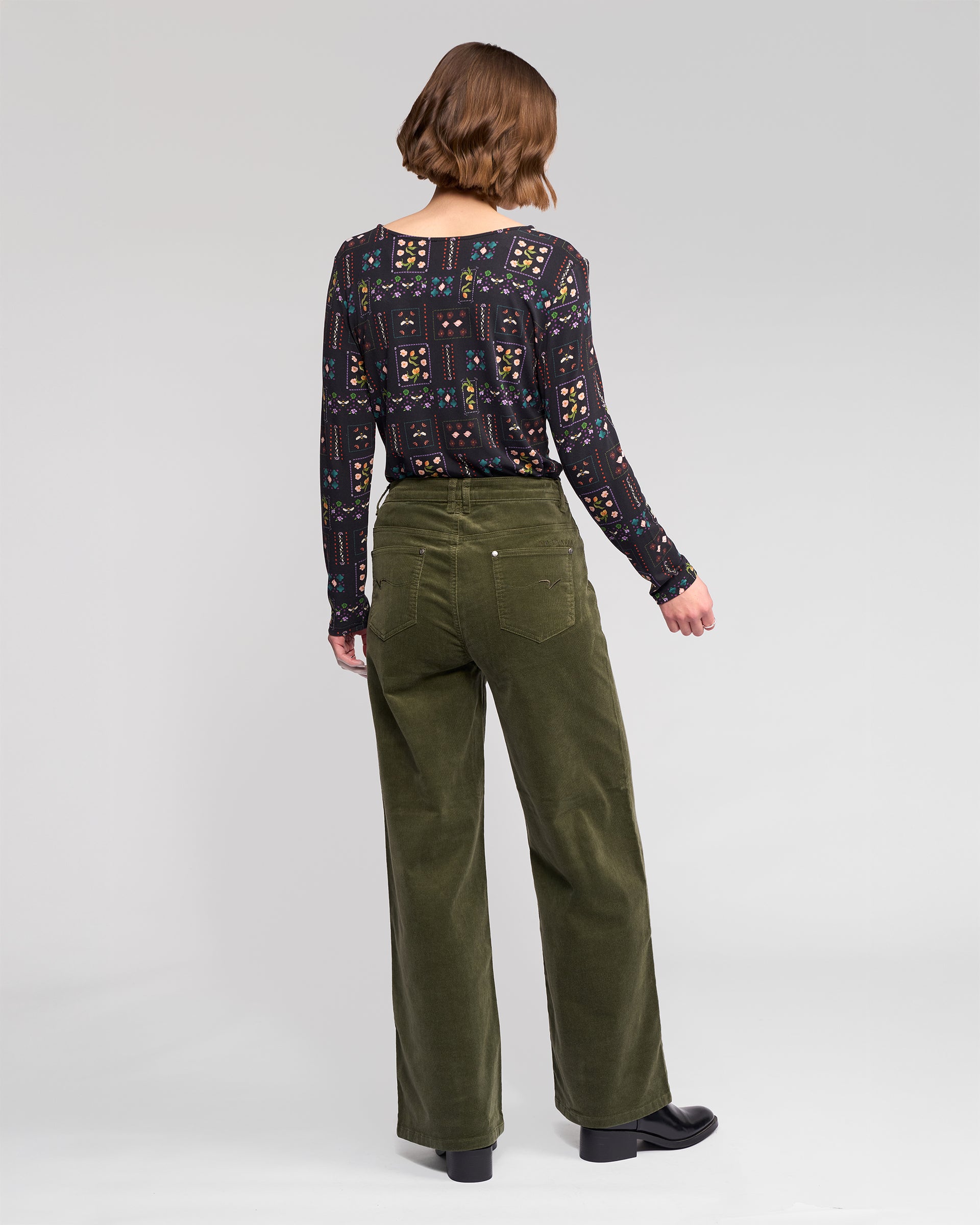 A person faces away, wearing Vassalli's Empire Cord Pant in Deep Sage with an indigo patterned long-sleeve top and black shoes against a plain light background.