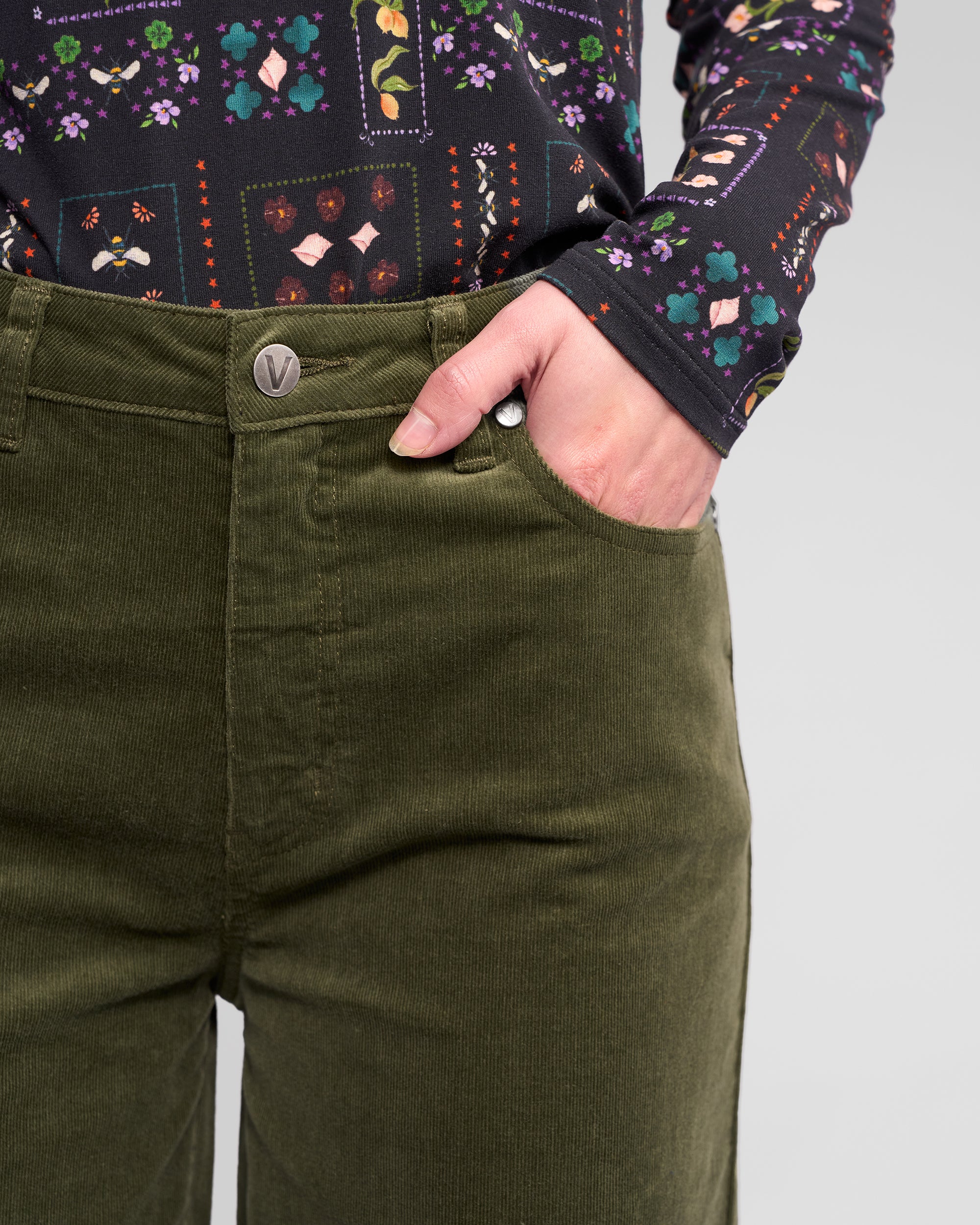 A person wears the Vassalli Empire Cord Pant in Deep Sage with a long-sleeve, colorful floral geometric shirt, one hand in their pocket, showcasing a chic winter look.