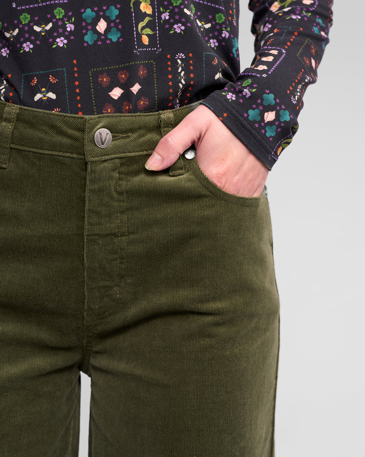A person wears the Vassalli Empire Cord Pant in Deep Sage with a long-sleeve, colorful floral geometric shirt, one hand in their pocket, showcasing a chic winter look.