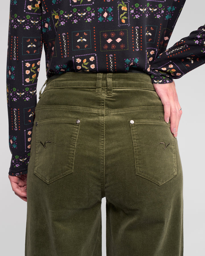 Wearing Vassalli's Empire Cord Pant in Deep Sage and a long-sleeved, patterned floral-geometric shirt, the person is shown from the back with one hand on their hip in a winter-ready look.