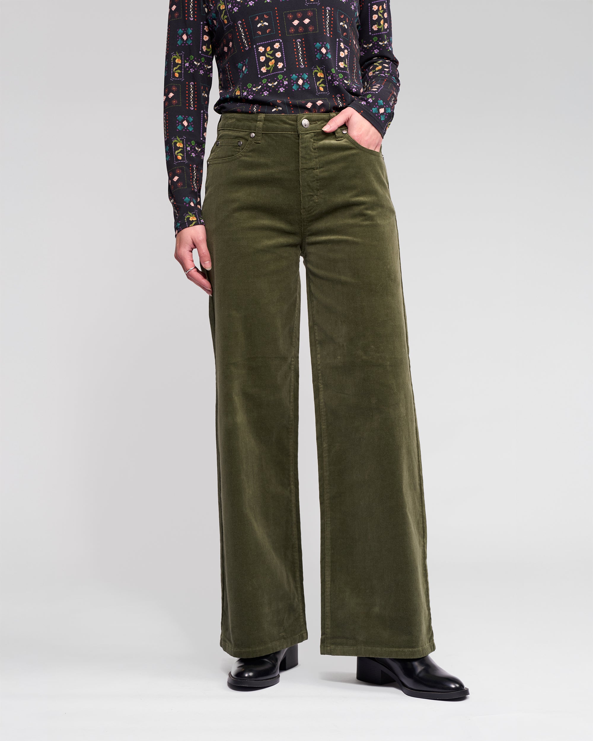 A person models the Vassalli Empire Cord Pant in Deep Sage with a patterned long-sleeve top and black heeled boots, shown from shoulder to feet against a light background for a winter-ready look.