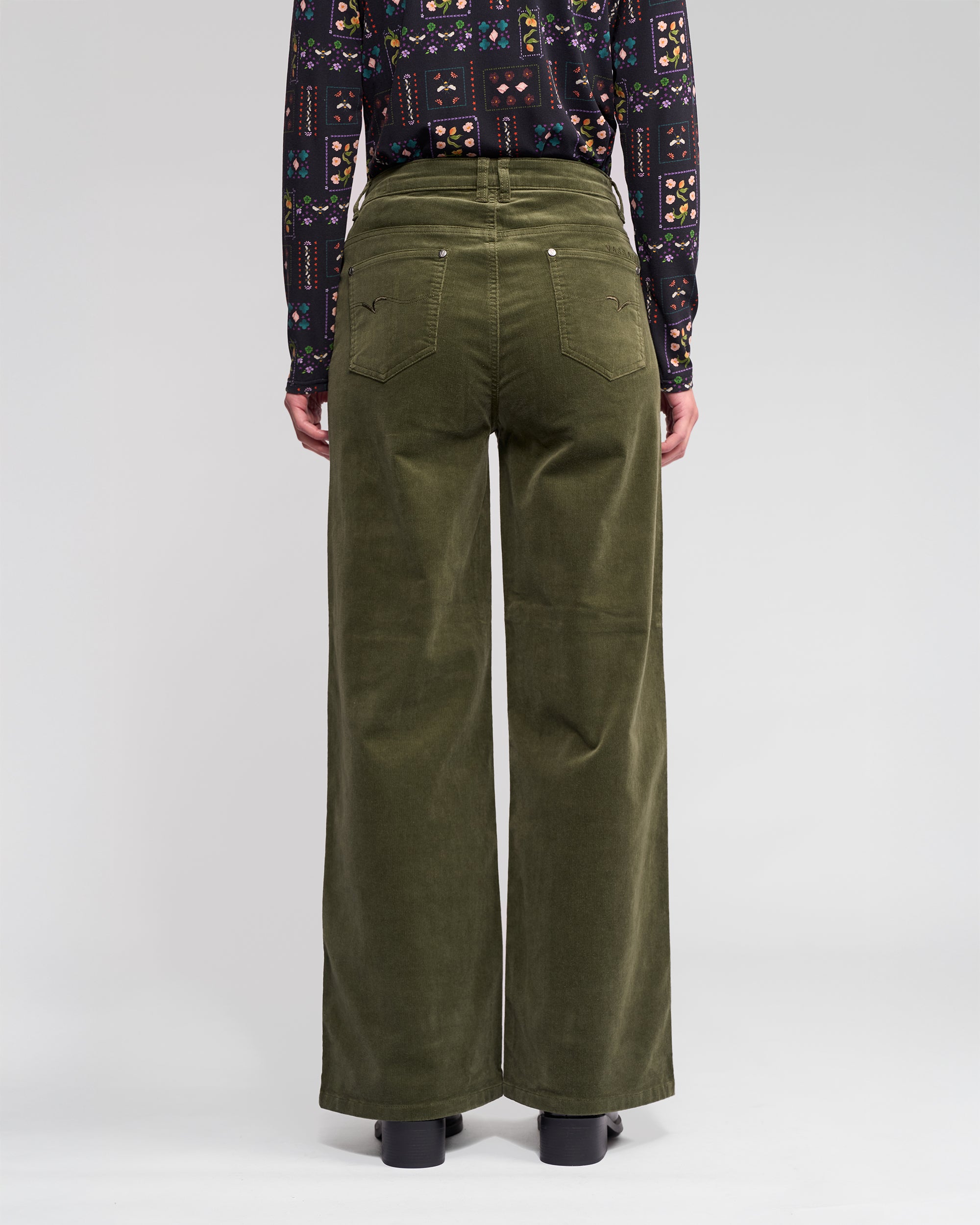 A person faces away, wearing Vassalli’s Empire Cord Pant in Deep Sage with back pockets, paired with a patterned long-sleeve shirt and black shoes against a plain light background.