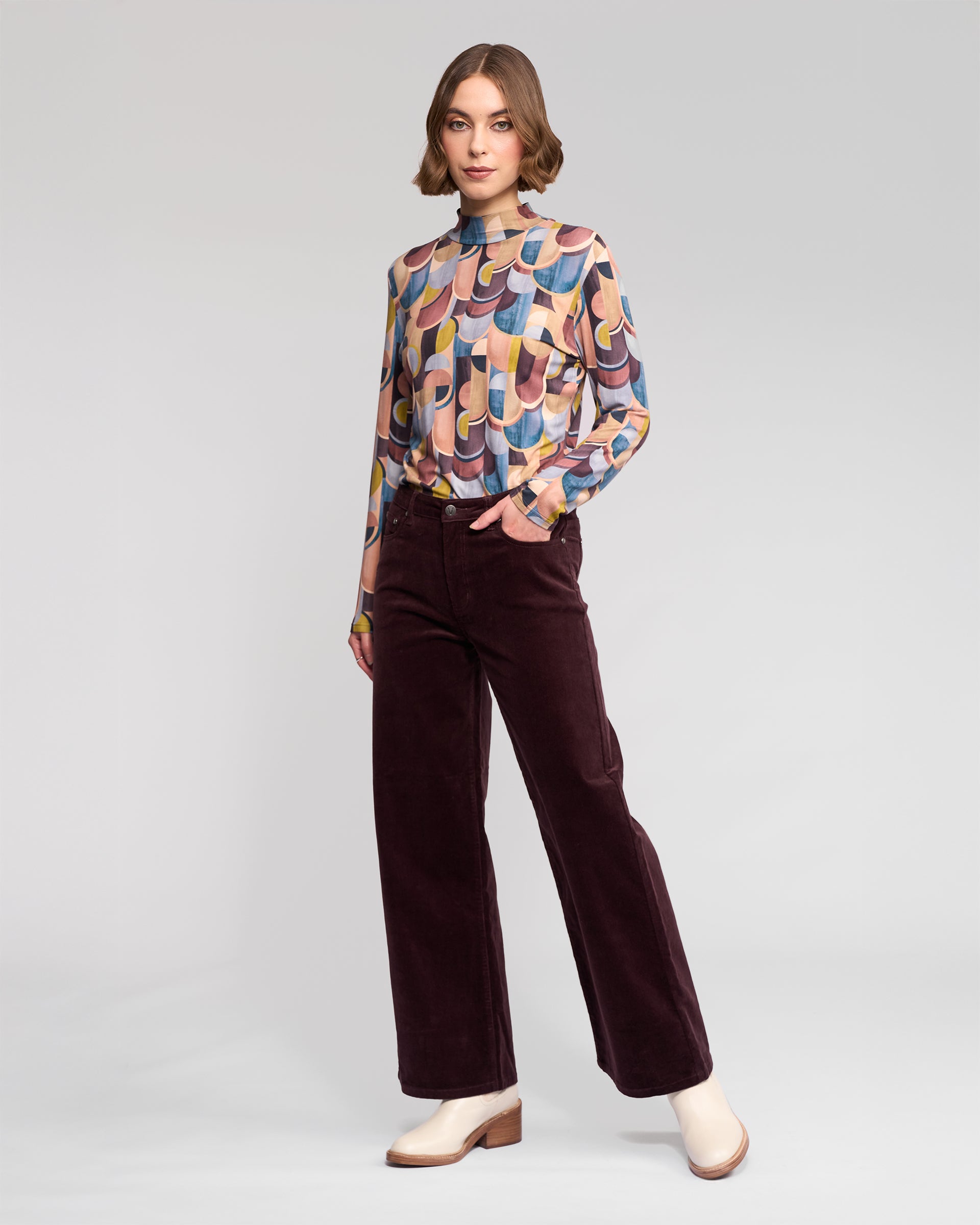A person poses confidently in a studio, wearing Vassalli’s Empire Cord Pant in Merlot paired with a colorful patterned blouse and light heeled shoes, with one hand in their pocket.