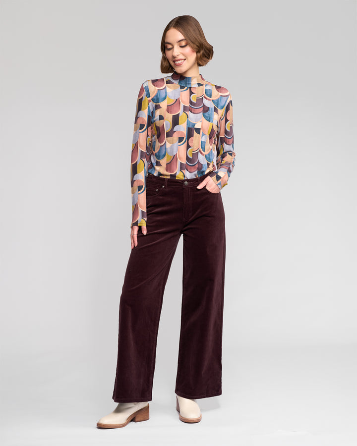 A woman stands against a plain background, smiling in a long-sleeve geometric-print top and Vassalli’s Empire Cord Pant in rich Merlot, paired with cream ankle boots and one hand casually in her pocket.