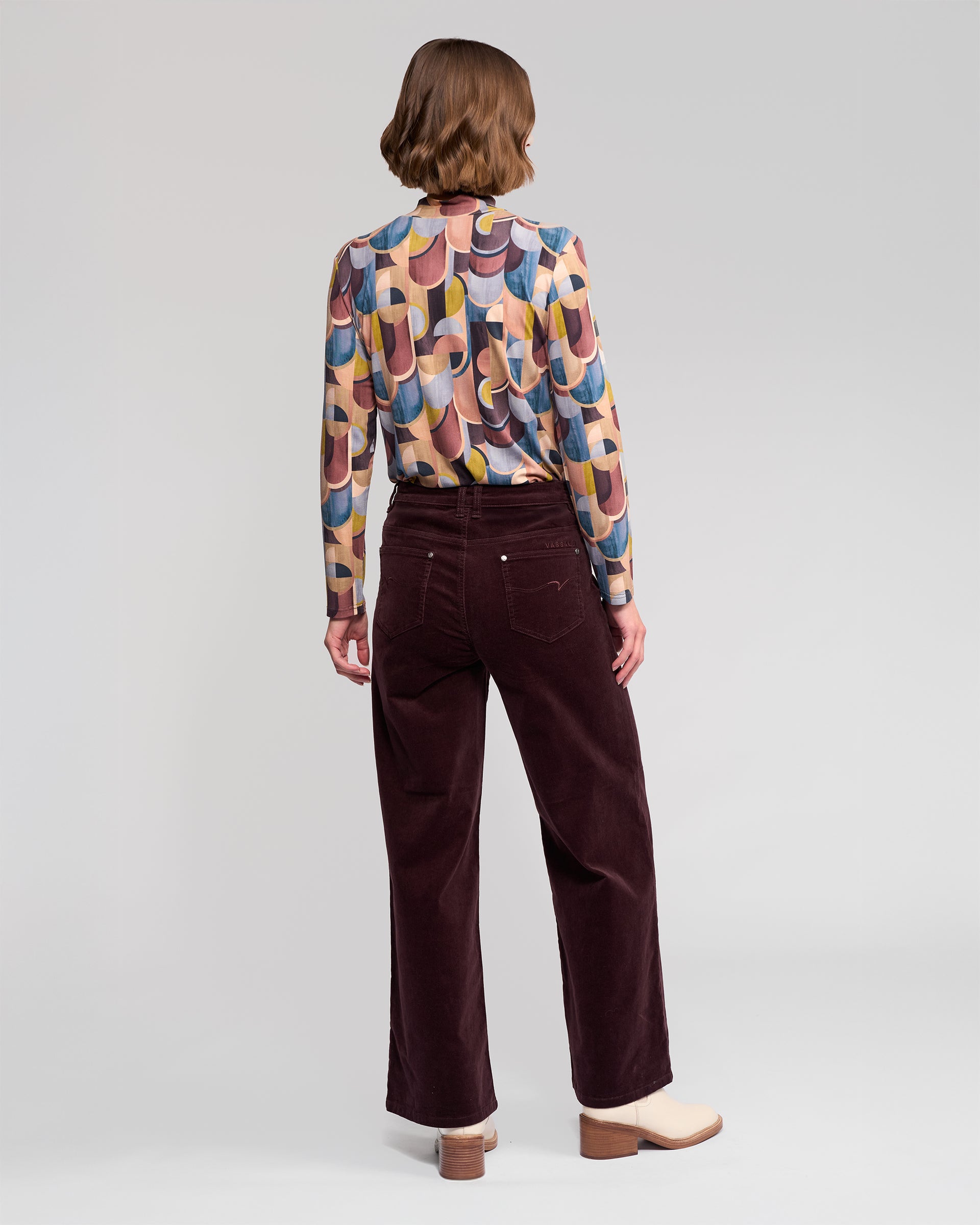 A person with short brown hair faces away, wearing a colorful patterned long-sleeve shirt, Vassalli's Empire Cord Pant in Merlot, and cream-colored heeled shoes against a plain light background.