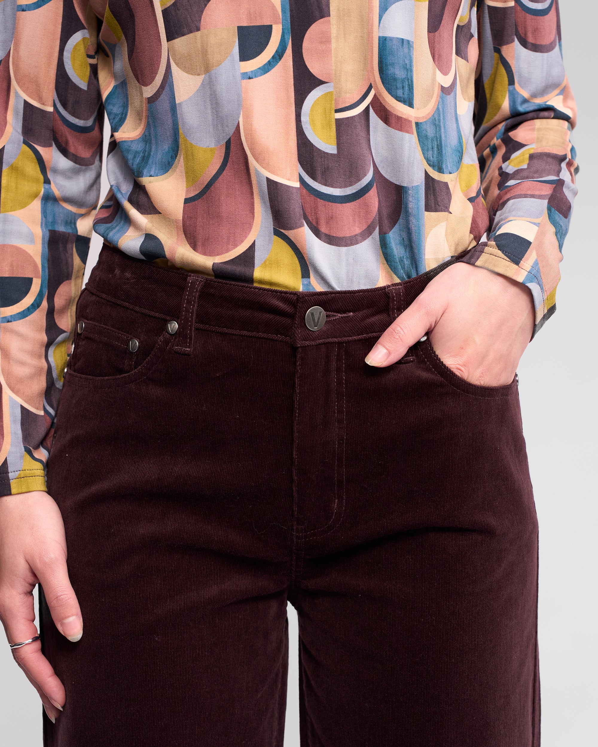 A person stands against a plain background, wearing Vassalli’s Empire Cord Pant in Merlot and a colorful shirt with curved patterns, one hand casually in their pocket.