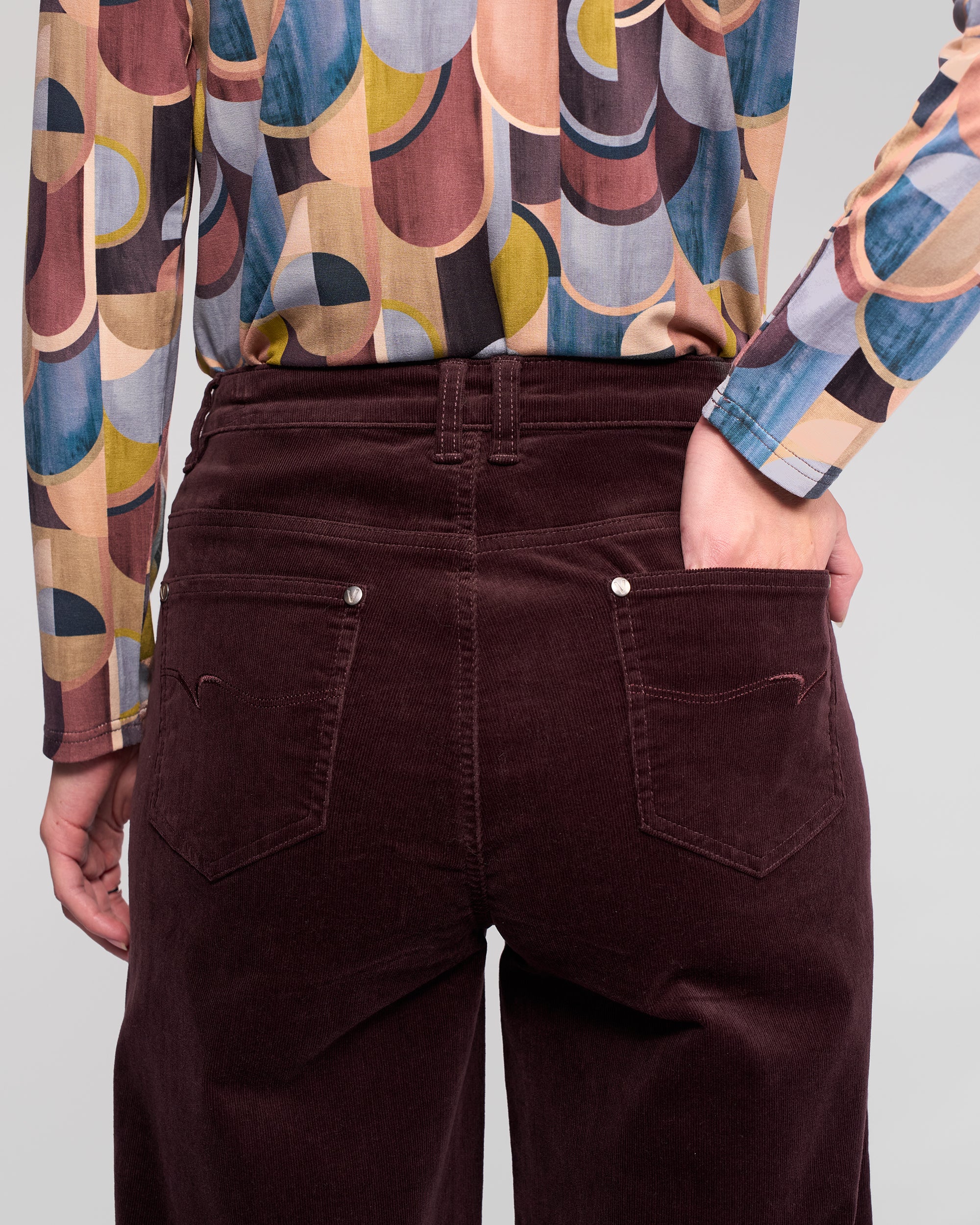 A person stands with one hand in the back pocket of their Vassalli Empire Cord Pant in Merlot, paired with a colorful geometric shirt. The rear-view image highlights the pant’s waist and upper legs, showcasing its winter-ready wide-leg style.