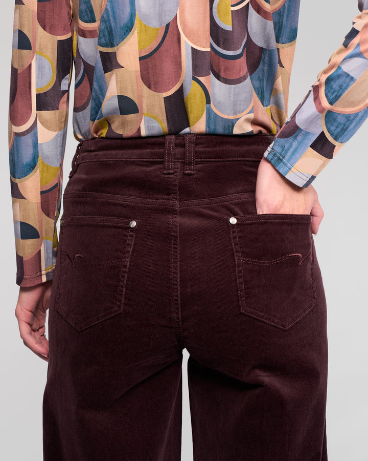 A person stands with one hand in the back pocket of their Vassalli Empire Cord Pant in Merlot, paired with a colorful geometric shirt. The rear-view image highlights the pant’s waist and upper legs, showcasing its winter-ready wide-leg style.