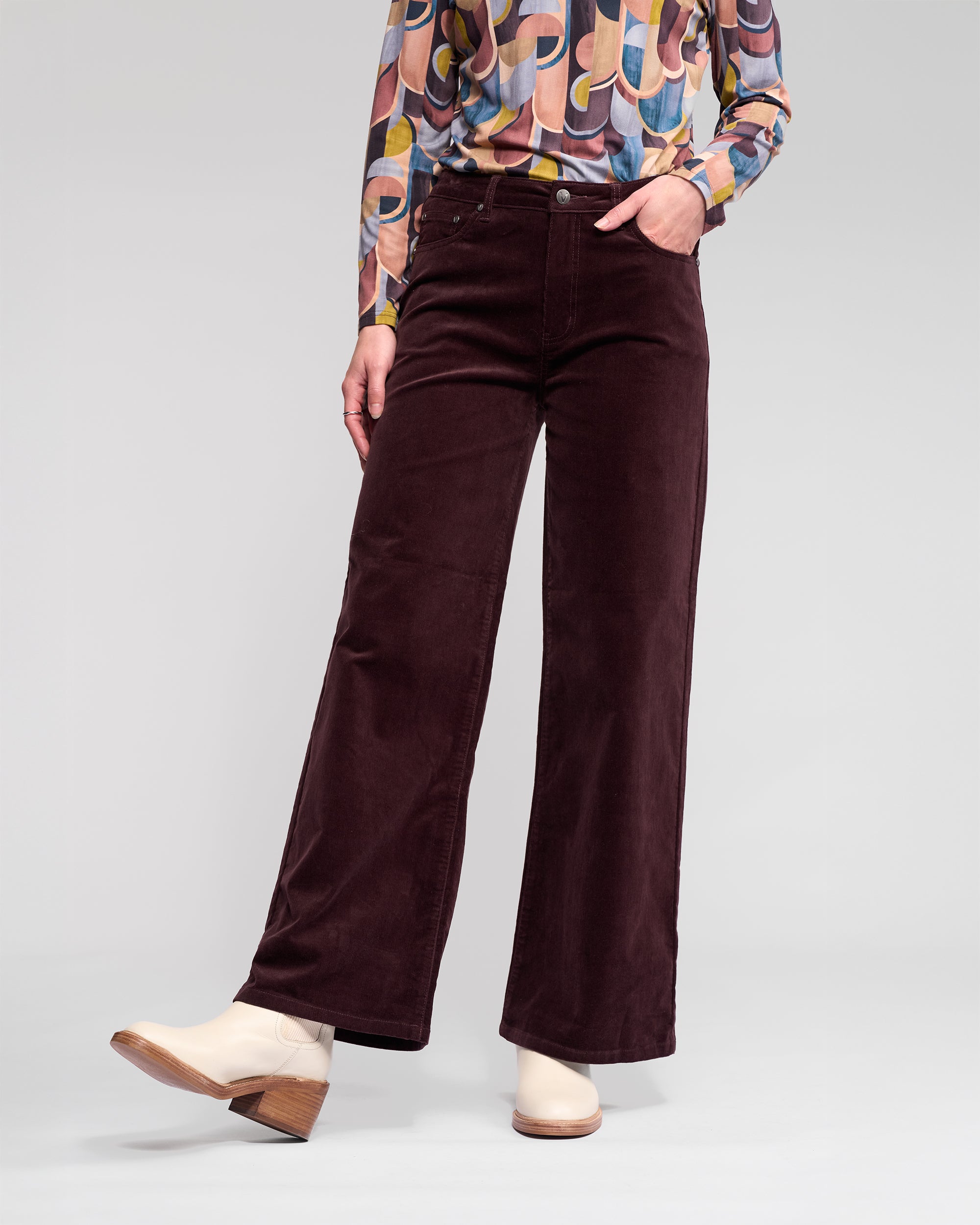 A person wears Vassalli's Empire Cord Pant in Merlot, paired with a colorful geometric blouse and off-white ankle boots, standing with one hand in their pocket against a plain background.