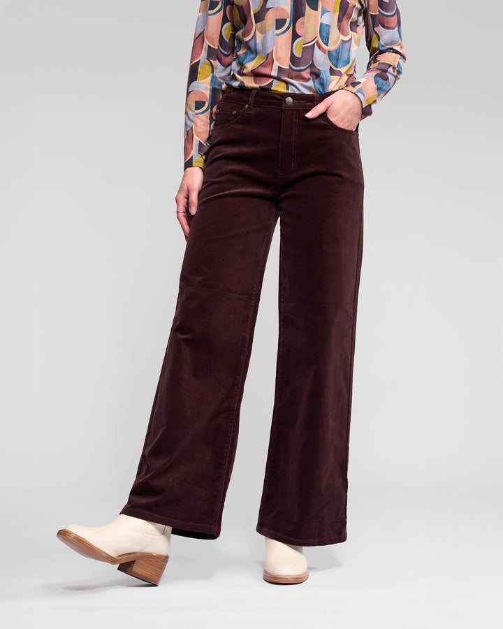 A person wears Vassalli's Empire Cord Pant in Merlot, paired with a colorful geometric blouse and off-white ankle boots, standing with one hand in their pocket against a plain background.