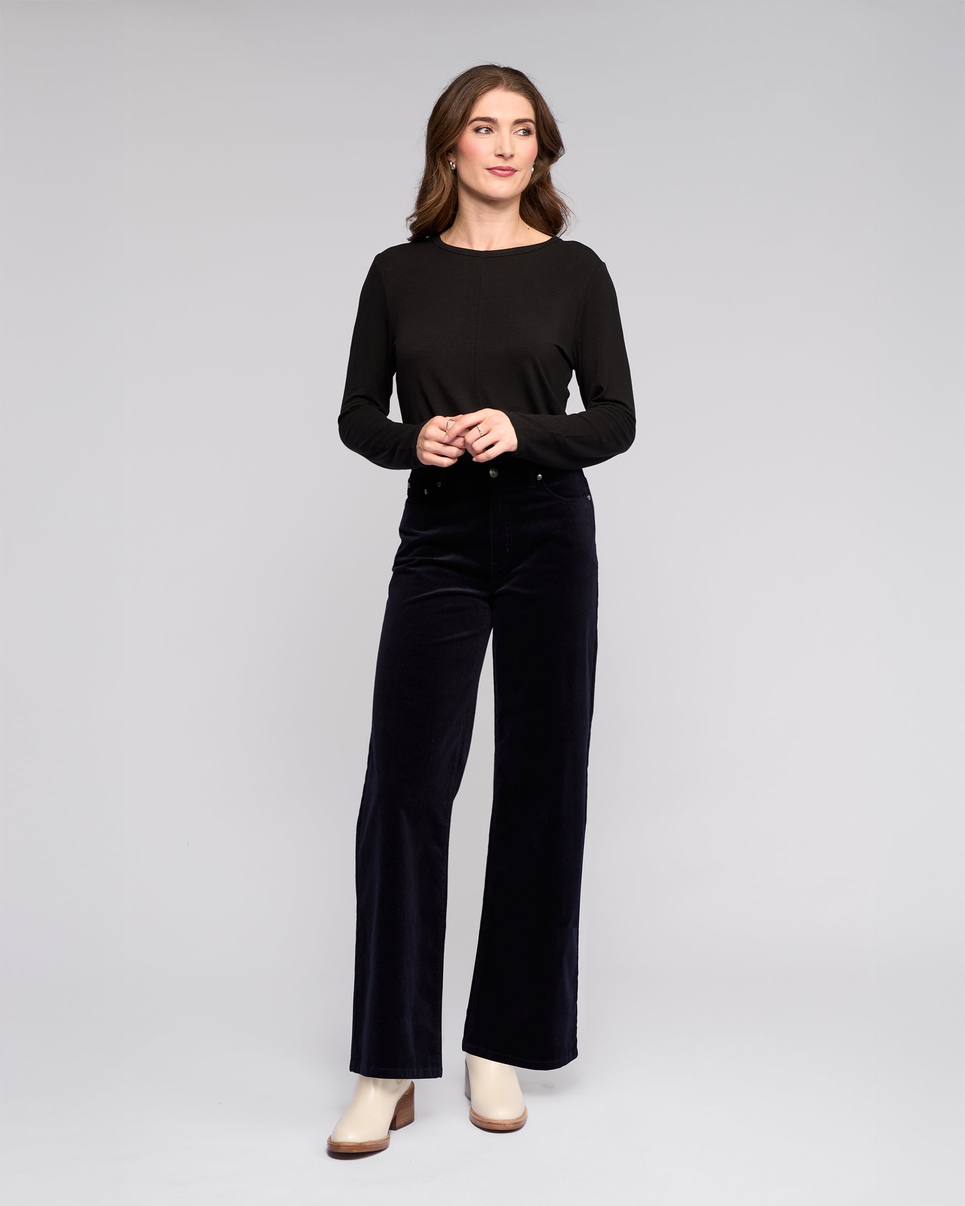 A woman stands against a light gray background, smiling softly as she looks to the side. She wears a long-sleeve black top, off-white ankle boots, and Vassalli's Empire Cord Pant in Midnight.