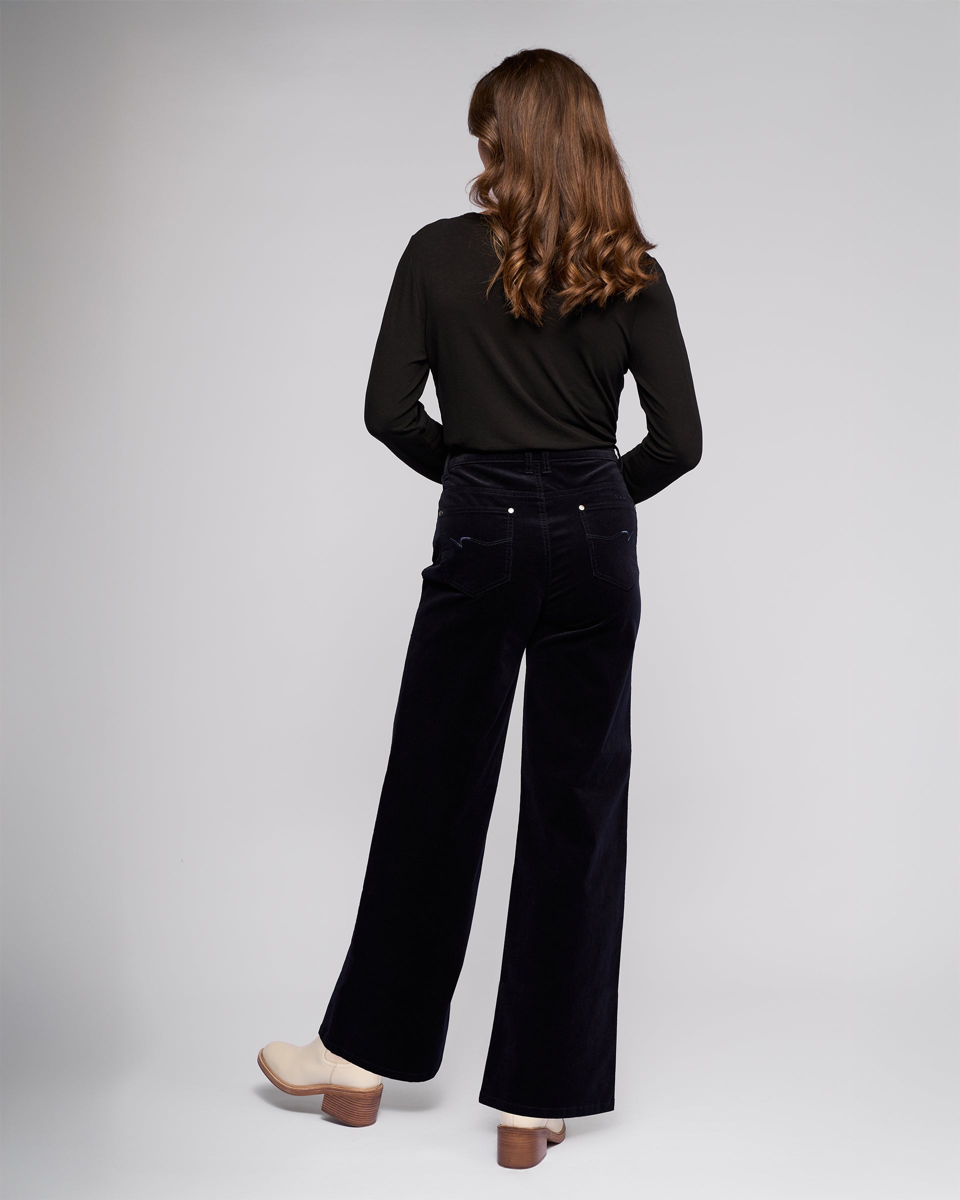 A woman with wavy brown hair models Vassalli’s Empire Cord Pant in Midnight, paired with a black long-sleeve top and beige heeled shoes, standing against a plain light gray background.