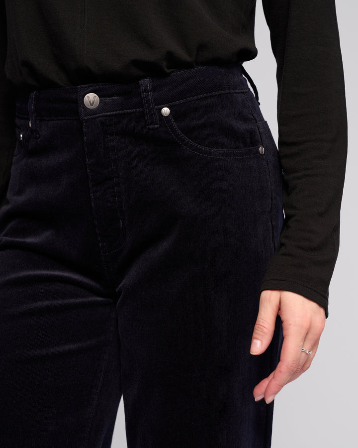 Wearing Vassalli's Empire Cord Pant in Midnight and a long-sleeved black top, a person stands with one hand by their side, subtly displaying a silver ring on their finger.
