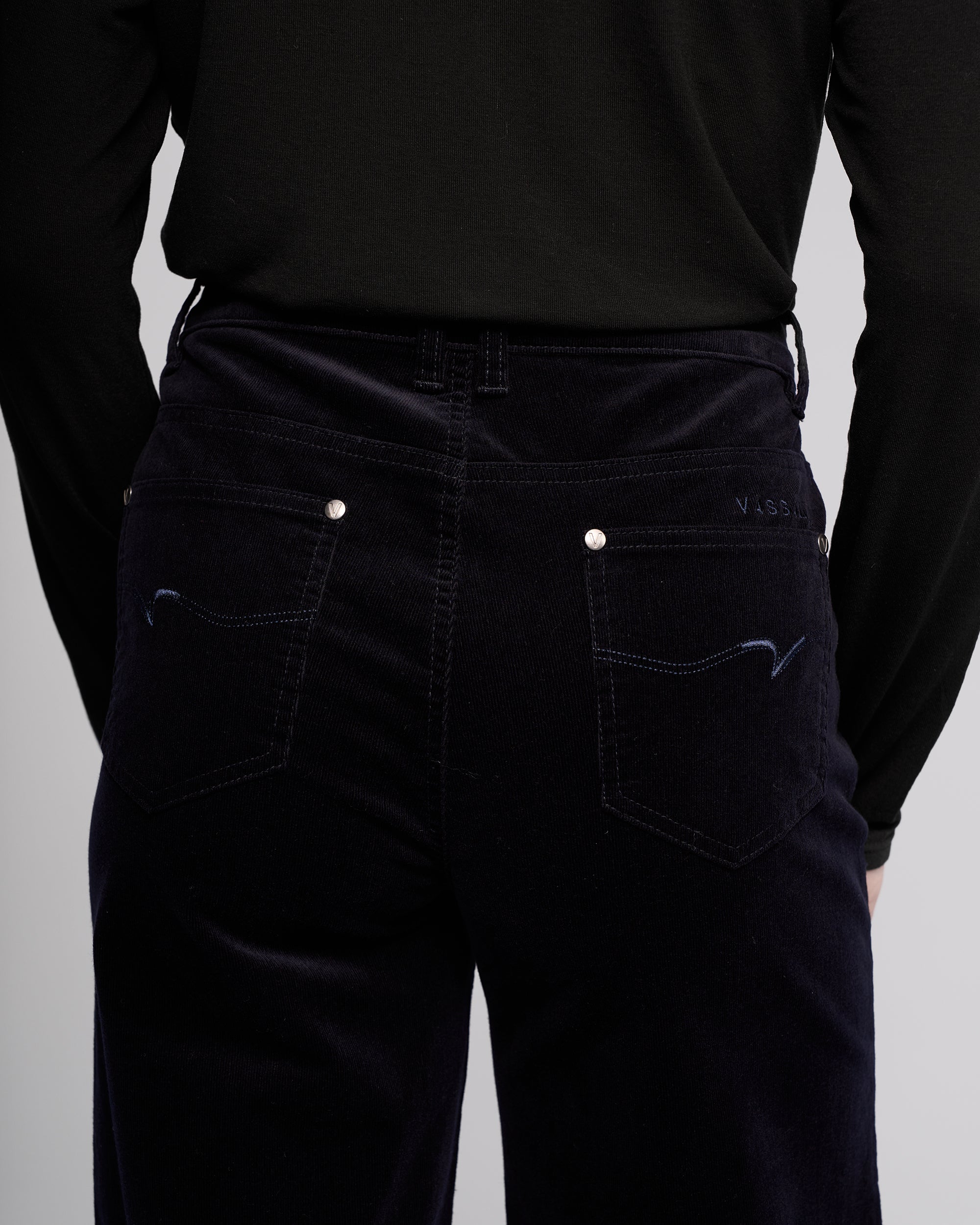 A person is shown from the back wearing Vassalli's Empire Cord Pant in Midnight, spotlighting the waist and upper legs. The winter-ready pants feature subtle stitching, silver rivets, and stylish back pockets.