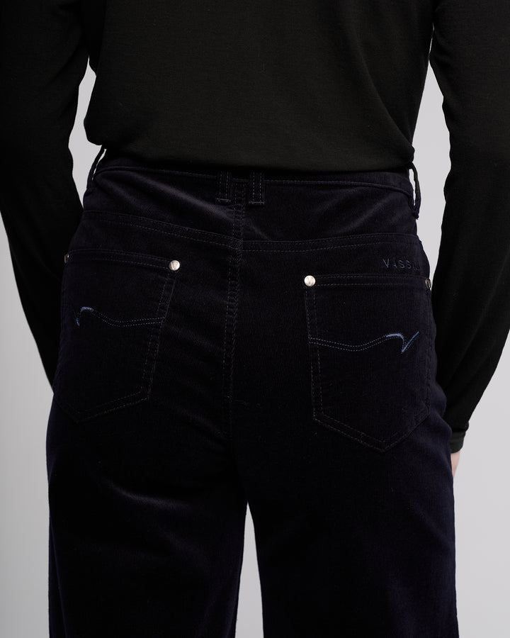 A person is shown from the back wearing Vassalli's Empire Cord Pant in Midnight, spotlighting the waist and upper legs. The winter-ready pants feature subtle stitching, silver rivets, and stylish back pockets.