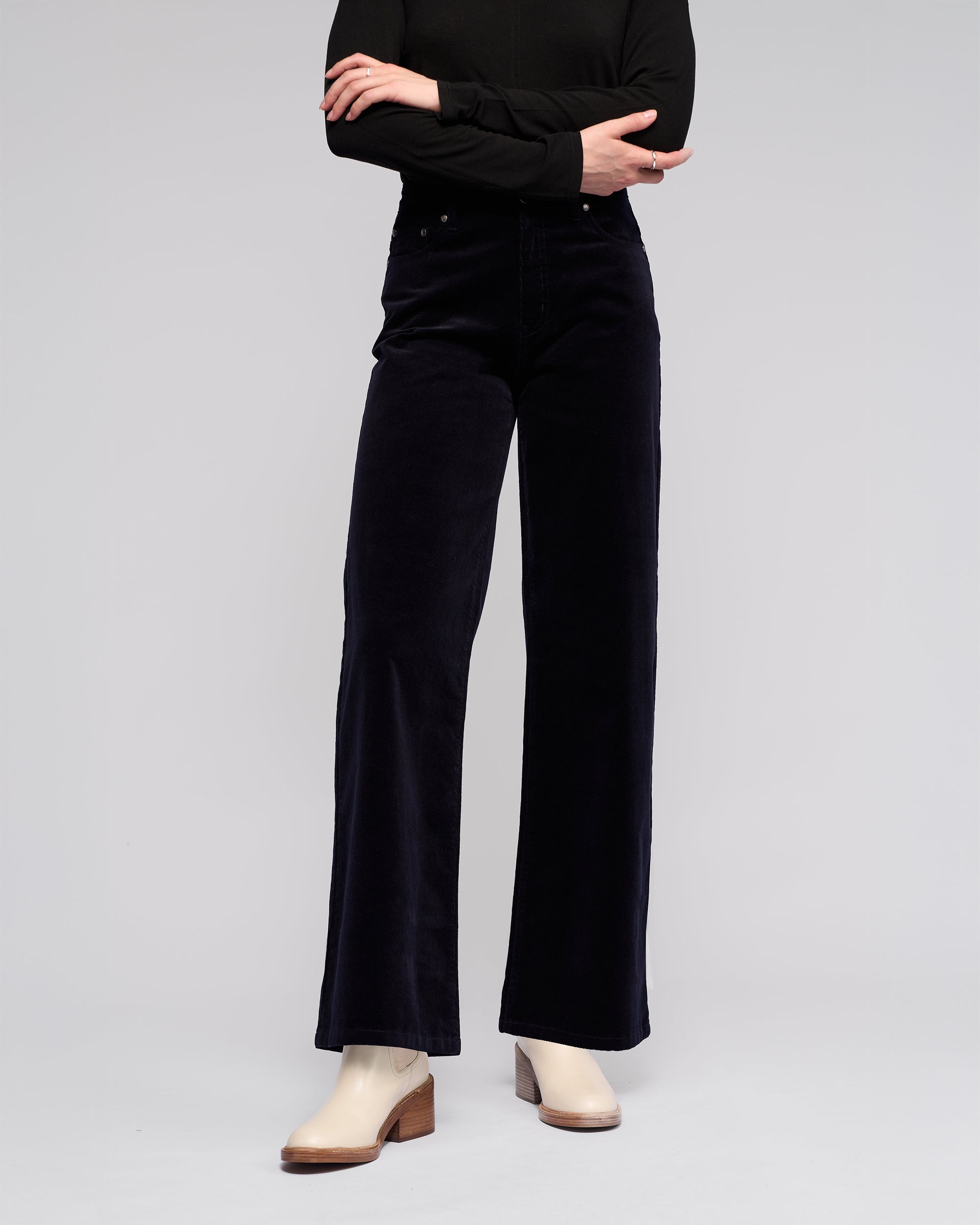 A person stands with arms crossed wearing a winter-ready black long-sleeve top, Vassalli Empire Cord Pant in Midnight, and beige low-heel ankle boots against a plain light background. Their head is not visible.