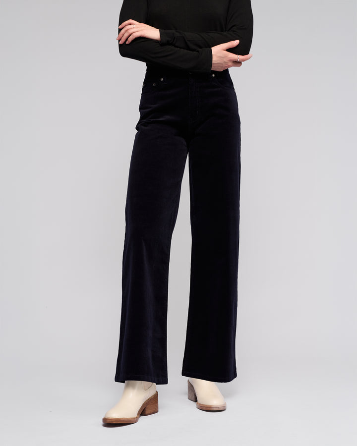 A person stands with arms crossed wearing a winter-ready black long-sleeve top, Vassalli Empire Cord Pant in Midnight, and beige low-heel ankle boots against a plain light background. Their head is not visible.