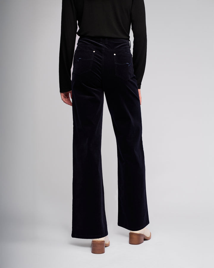 A person faces away, showing the back of their winter-ready look in a black long-sleeve top and Vassalli’s Empire Cord Pant in Midnight. The outfit is finished with light-colored heeled shoes.