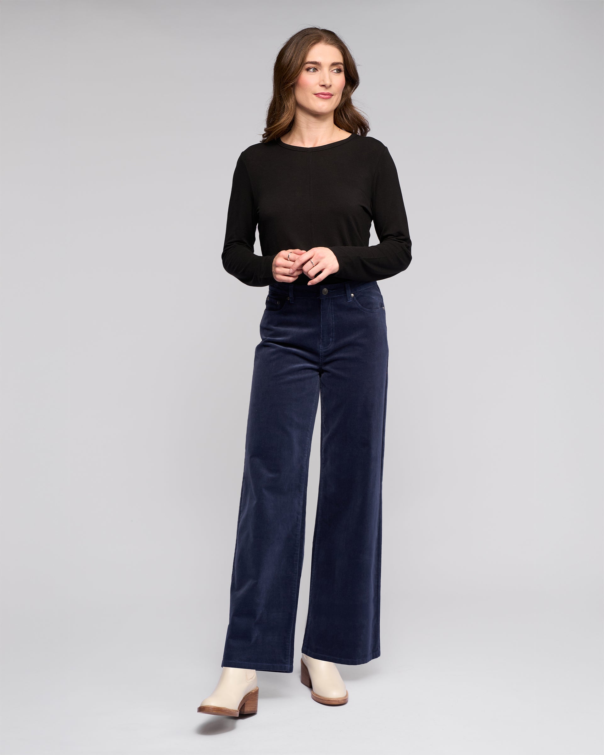 A woman with long brown hair and a neutral expression wears a black long-sleeve top, beige heeled shoes, and the Vassalli Empire Cord Pant in Slate, standing against a plain background.