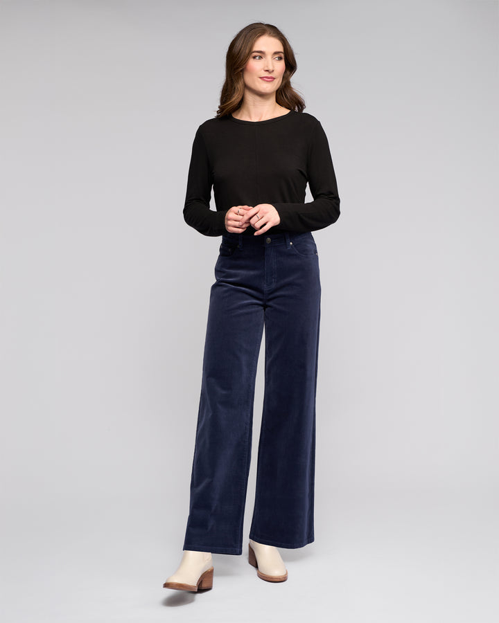 A woman with long brown hair and a neutral expression wears a black long-sleeve top, beige heeled shoes, and the Vassalli Empire Cord Pant in Slate, standing against a plain background.