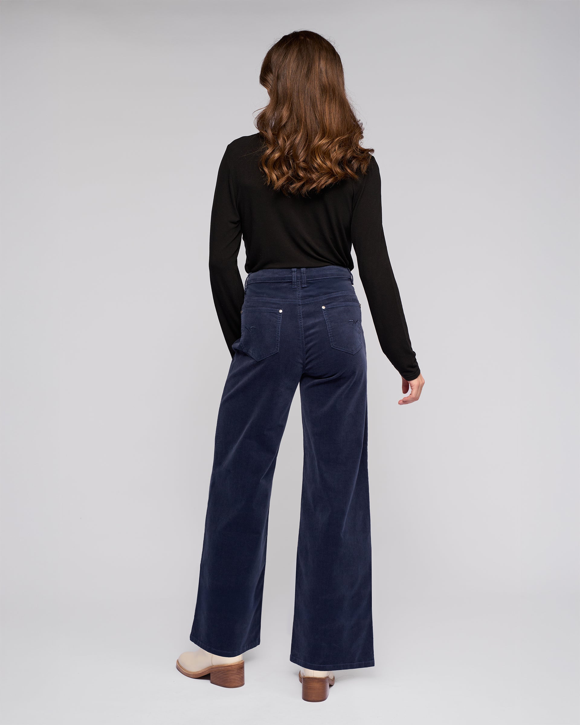 A person with long wavy brown hair, wearing a black long-sleeve top, Vassalli's Empire Cord Pant in Slate, and beige platform shoes stands facing away against a plain gray background.