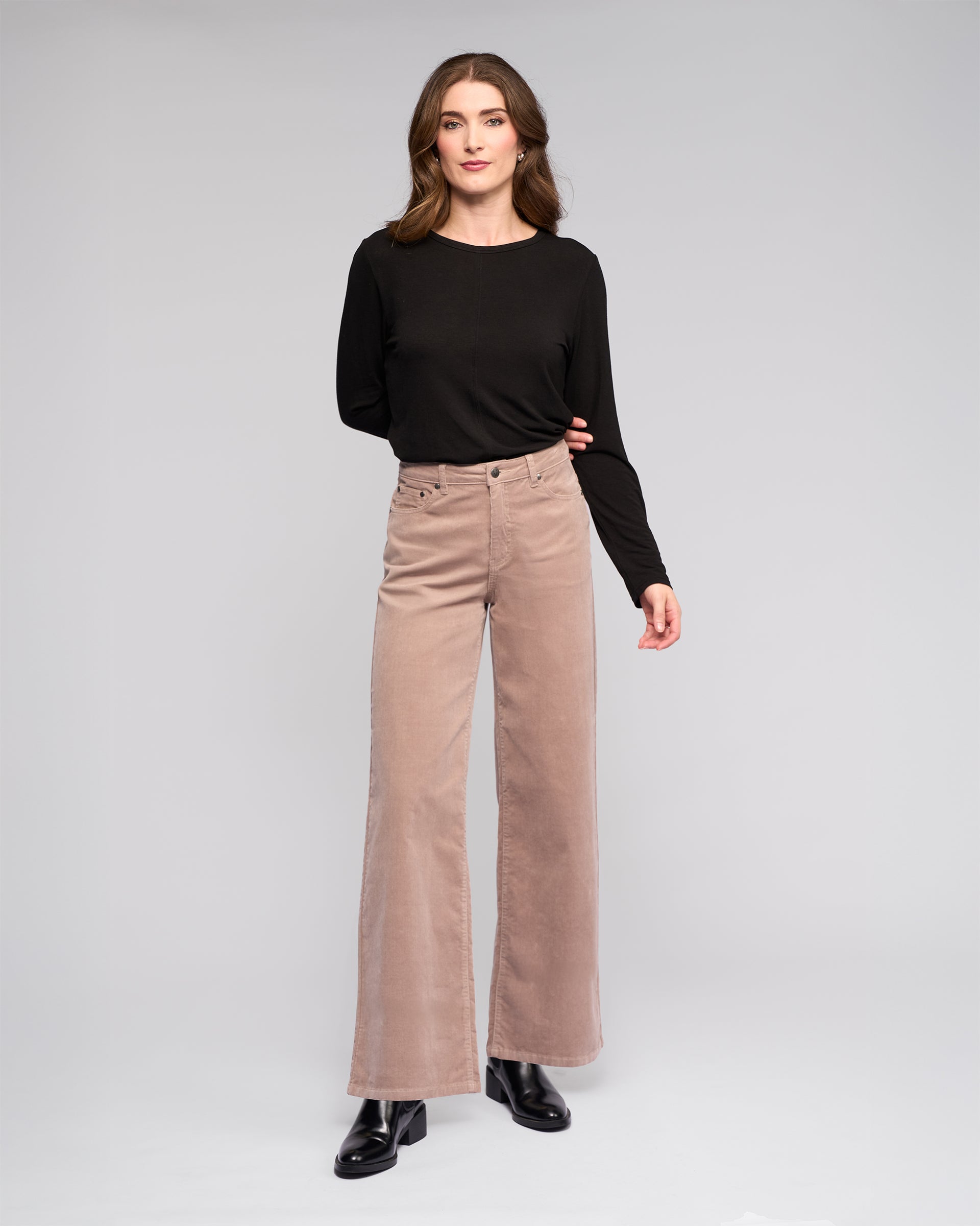 A woman with wavy brown hair and a neutral expression wears a black long-sleeve top, black ankle boots, and the Vassalli Empire Cord Pant in Twig against a plain background.