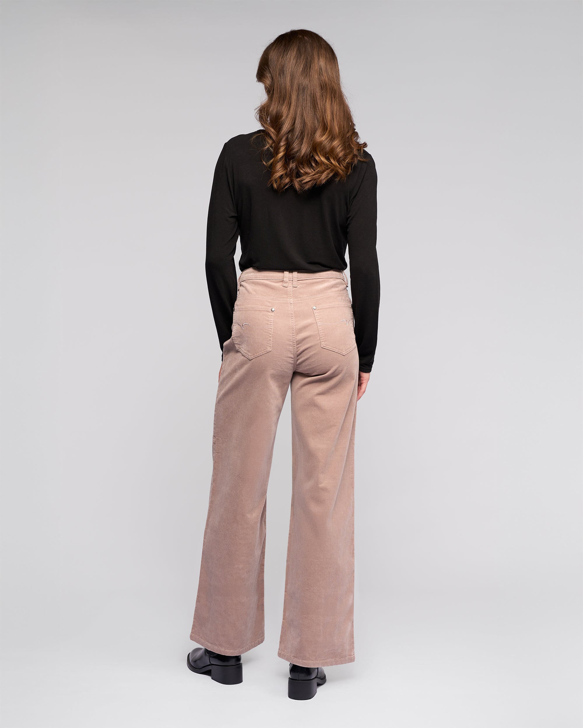 A woman with wavy brown hair faces away, wearing a Vassalli Empire Cord Pant in Twig and a black long-sleeve top with black shoes against a light gray background.