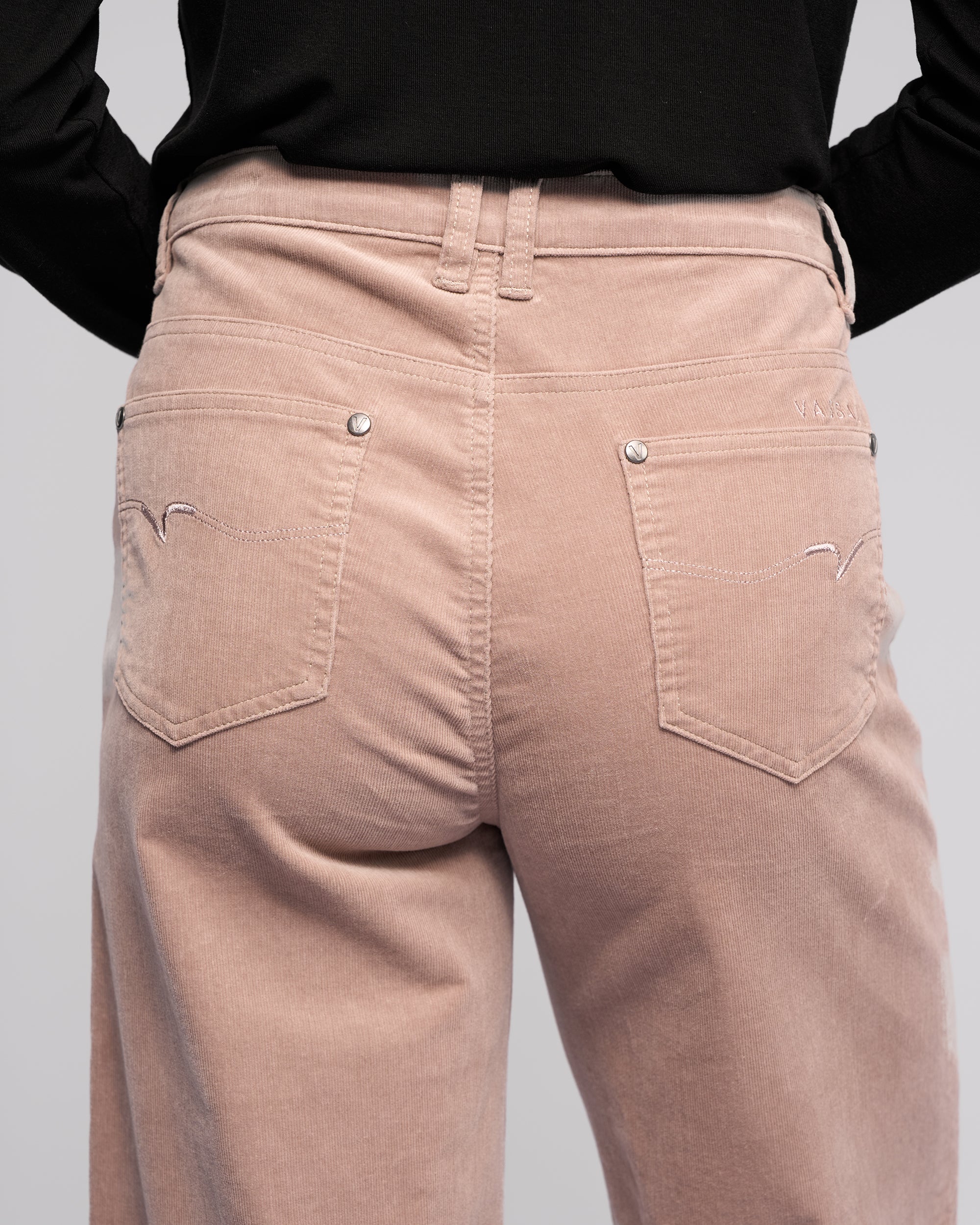 A person wearing Vassalli's Empire Cord Pant in Twig stands facing away, showcasing the winter-ready, wide-leg design along with back pocket and waistband details, paired with a black top.