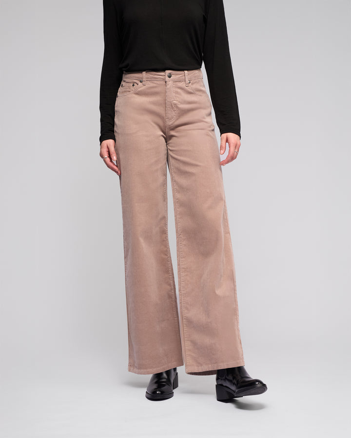 A person stands against a plain light background wearing a long-sleeve black top, black shoes, and Vassalli’s Empire Cord Pant in Twig. The image is cropped at the shoulders, so the face isn’t visible.