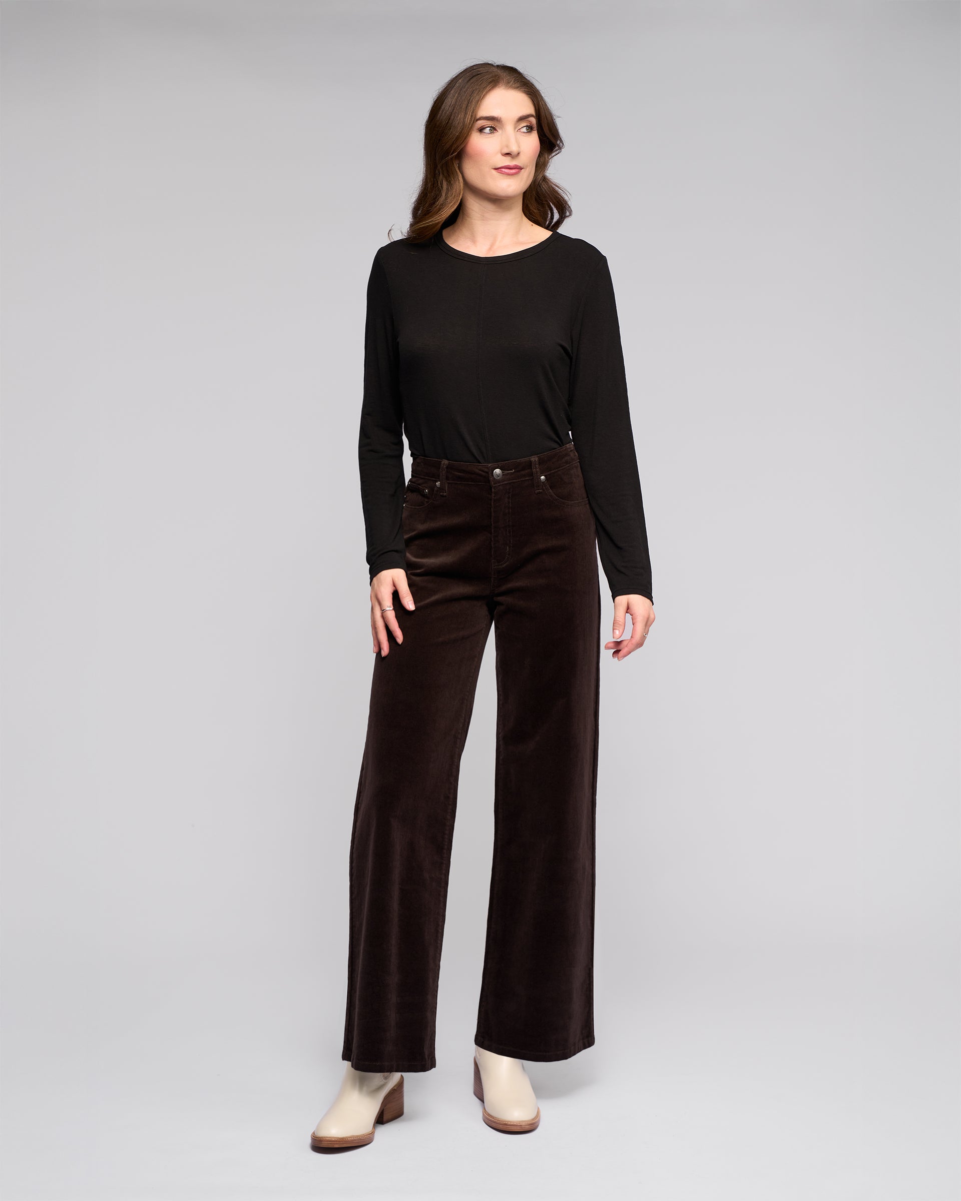 A woman with wavy brown hair stands against a plain background, wearing a long-sleeve black top, Vassalli's Empire Cord Pant in walnut, and light beige heeled boots. She looks slightly to the side.