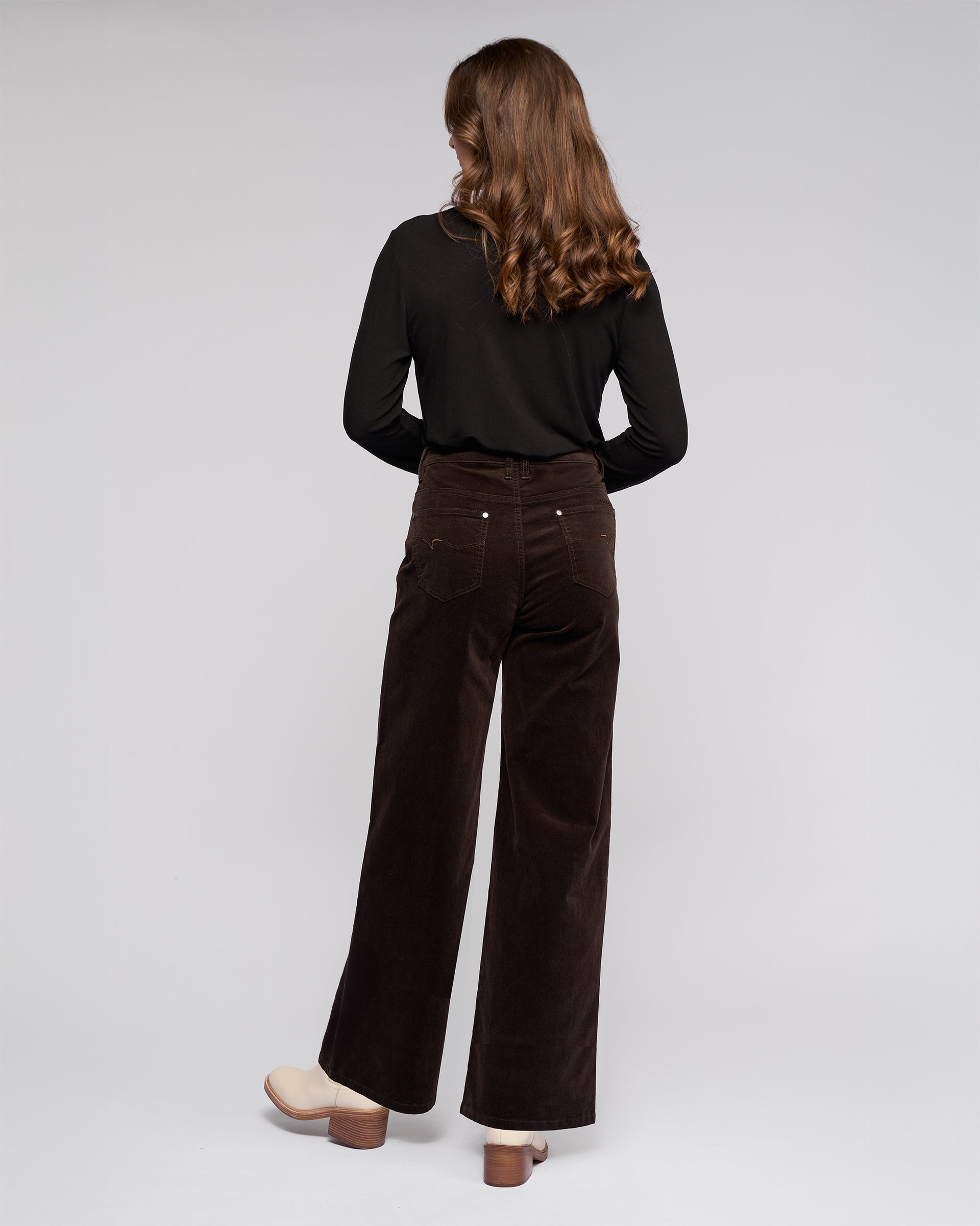 A woman with wavy brown hair, wearing a black long-sleeve top, beige heeled shoes, and Vassalli’s Empire Cord Pant in Walnut, stands facing away against a light background.