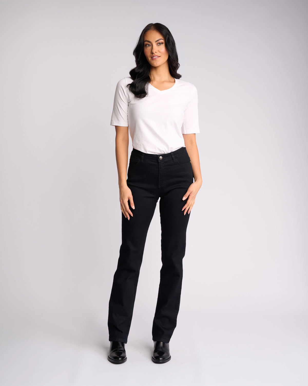 A woman with long dark hair wears the Vassalli Tour Jean in Black Denim, paired with a white short-sleeve t-shirt and black shoes. She stands against a plain background, facing forward with her right hand by her side and a neutral expression.