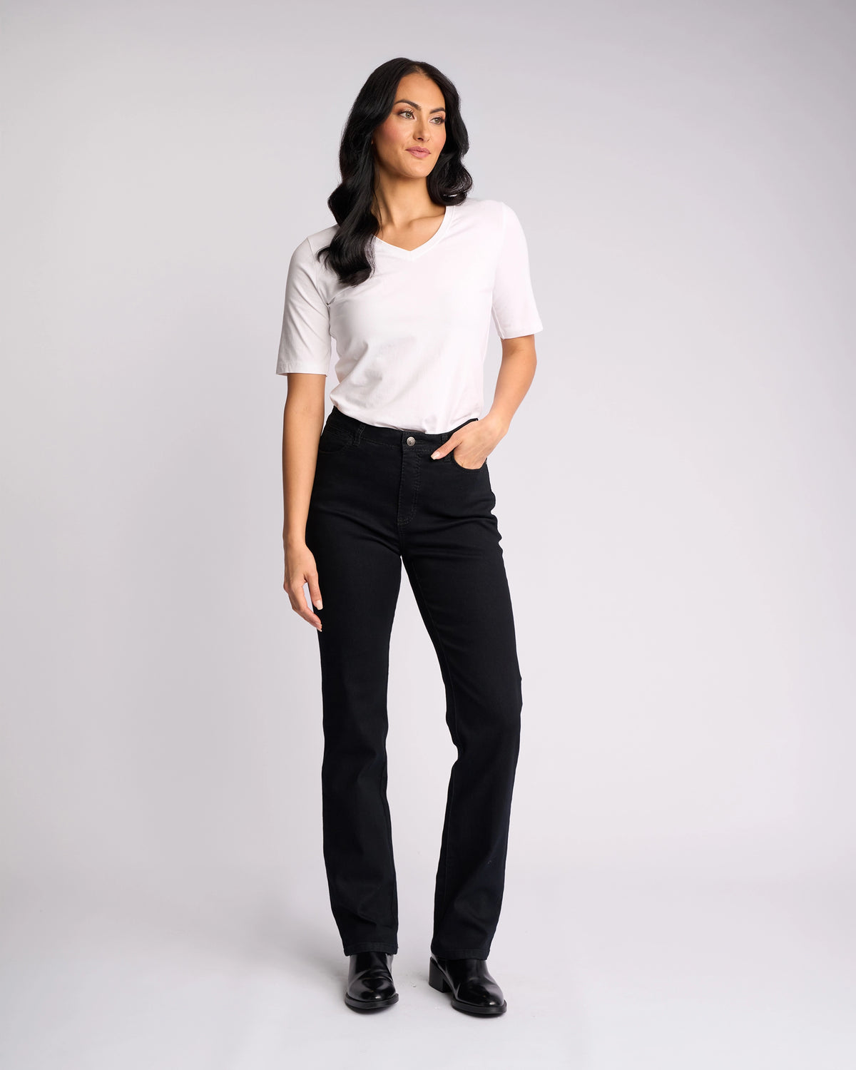 A woman with long dark hair stands against a light background, wearing a Vassalli Tour Jean in black denim with a high-rise waist and a white short-sleeve shirt. One hand is in her pocket; she completes the look with black shoes.