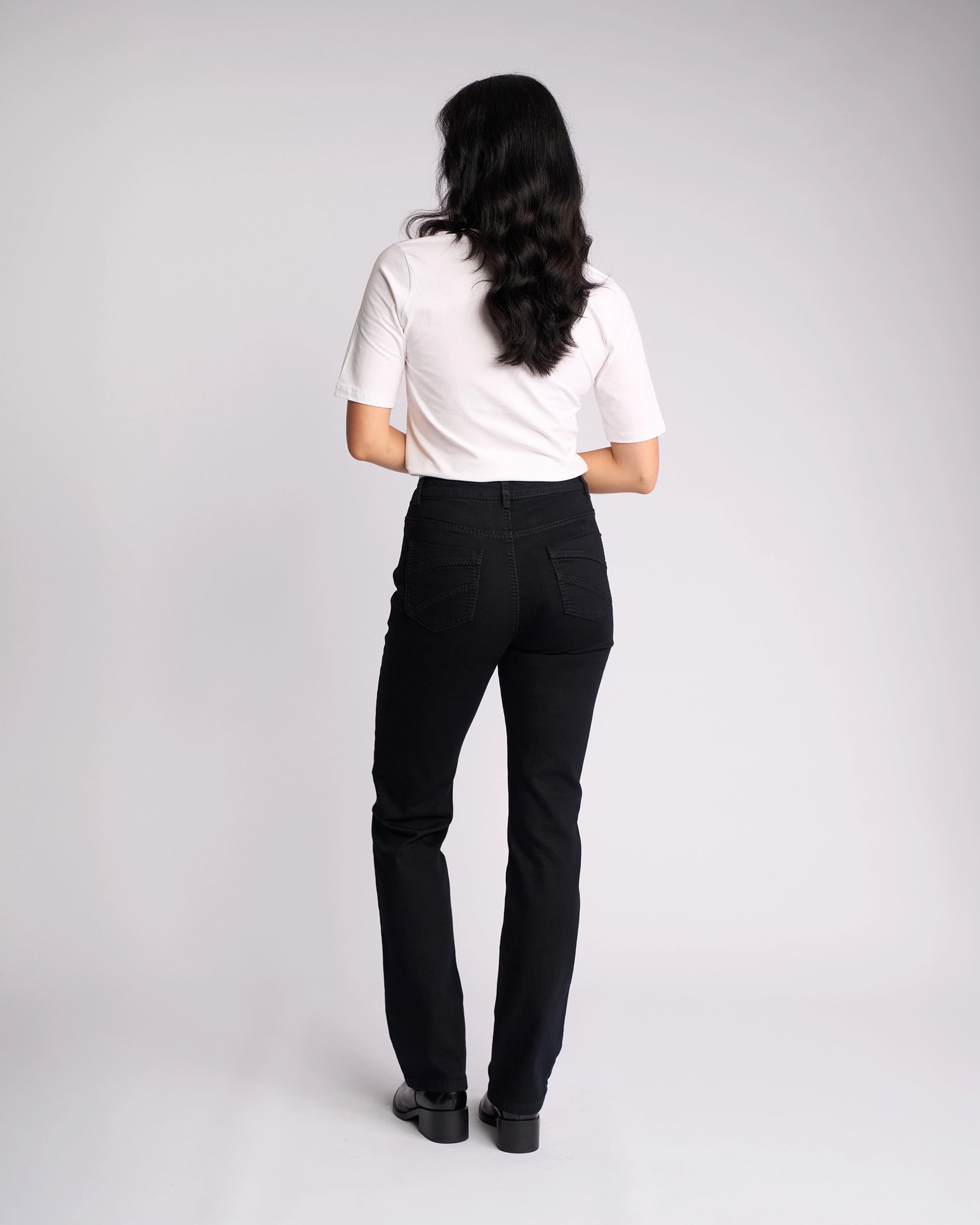 A woman with long dark hair wears a white short-sleeve top and Vassalli Tour Jean in Black Denim, featuring a high-rise waist, as she stands facing away from the camera against a plain light background.