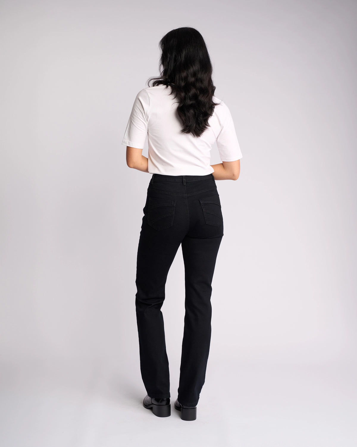 A woman with long dark hair wears a white short-sleeve top and Vassalli Tour Jean in Black Denim, featuring a high-rise waist, as she stands facing away from the camera against a plain light background.