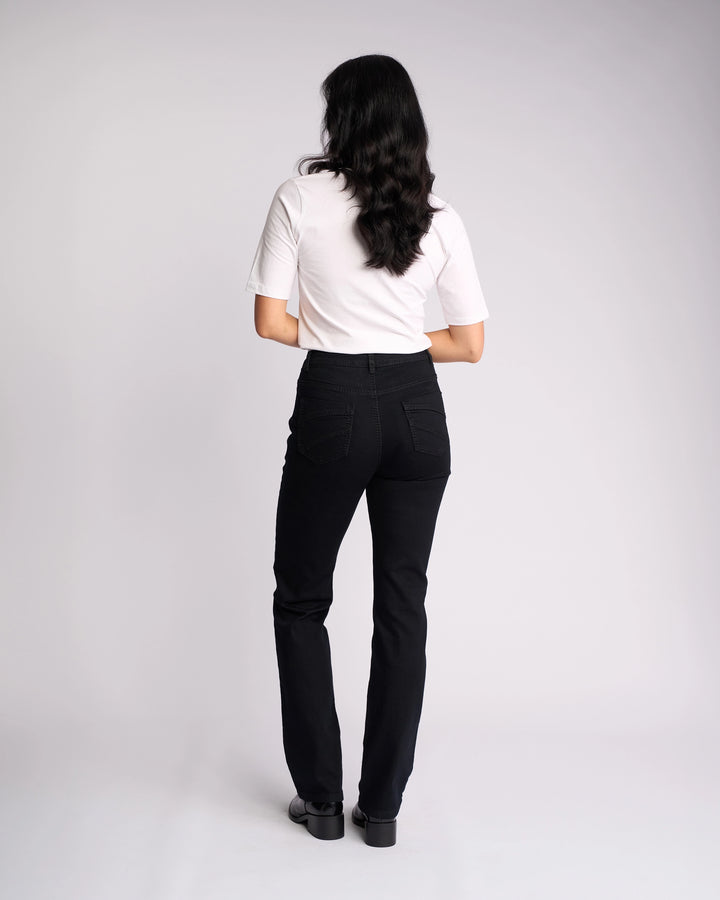 A woman with long dark hair wears a white short-sleeve top and Vassalli Tour Jean in Black Denim, featuring a high-rise waist, as she stands facing away from the camera against a plain light background.