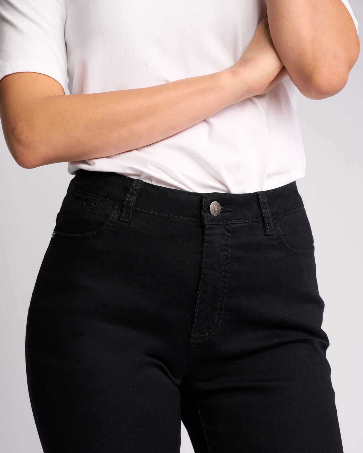 A person wearing a white short-sleeve shirt and Vassalli Tour Jean - Black Denim, featuring a high-rise waist, stands with one arm crossed and the other hand touching their face against a plain light background.