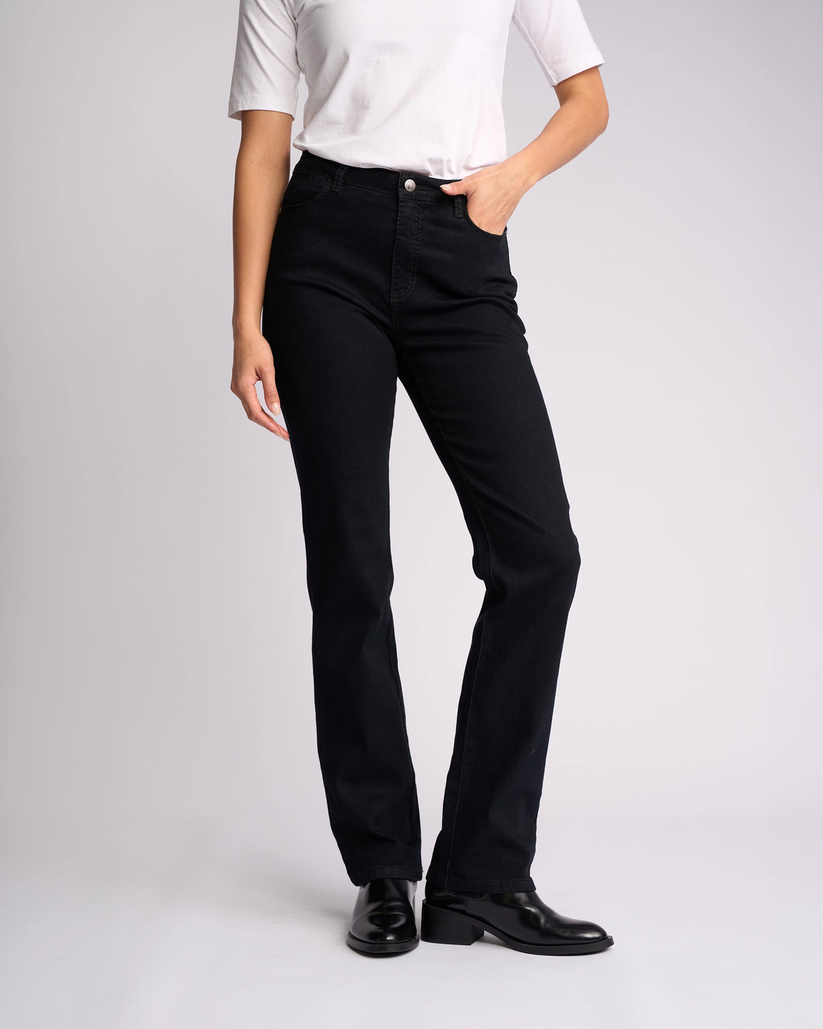 A person wearing a white short-sleeve top, Vassalli Tour Jean - Black Denim, and black shoes stands with one hand in their pocket against a plain light background. Their face is not visible.