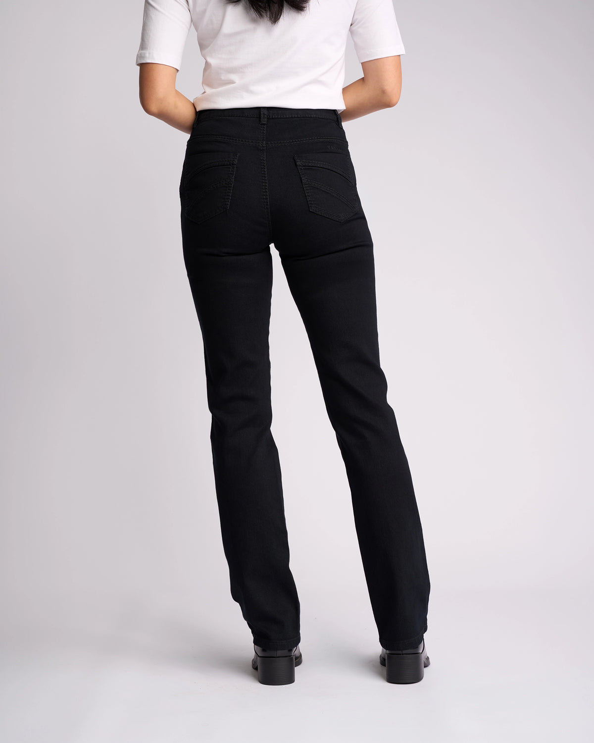 A woman faces away in a white short-sleeve top and Vassalli Tour Jean - Black Denim, featuring a high-rise waist and back pockets, paired with black shoes against a plain light background.