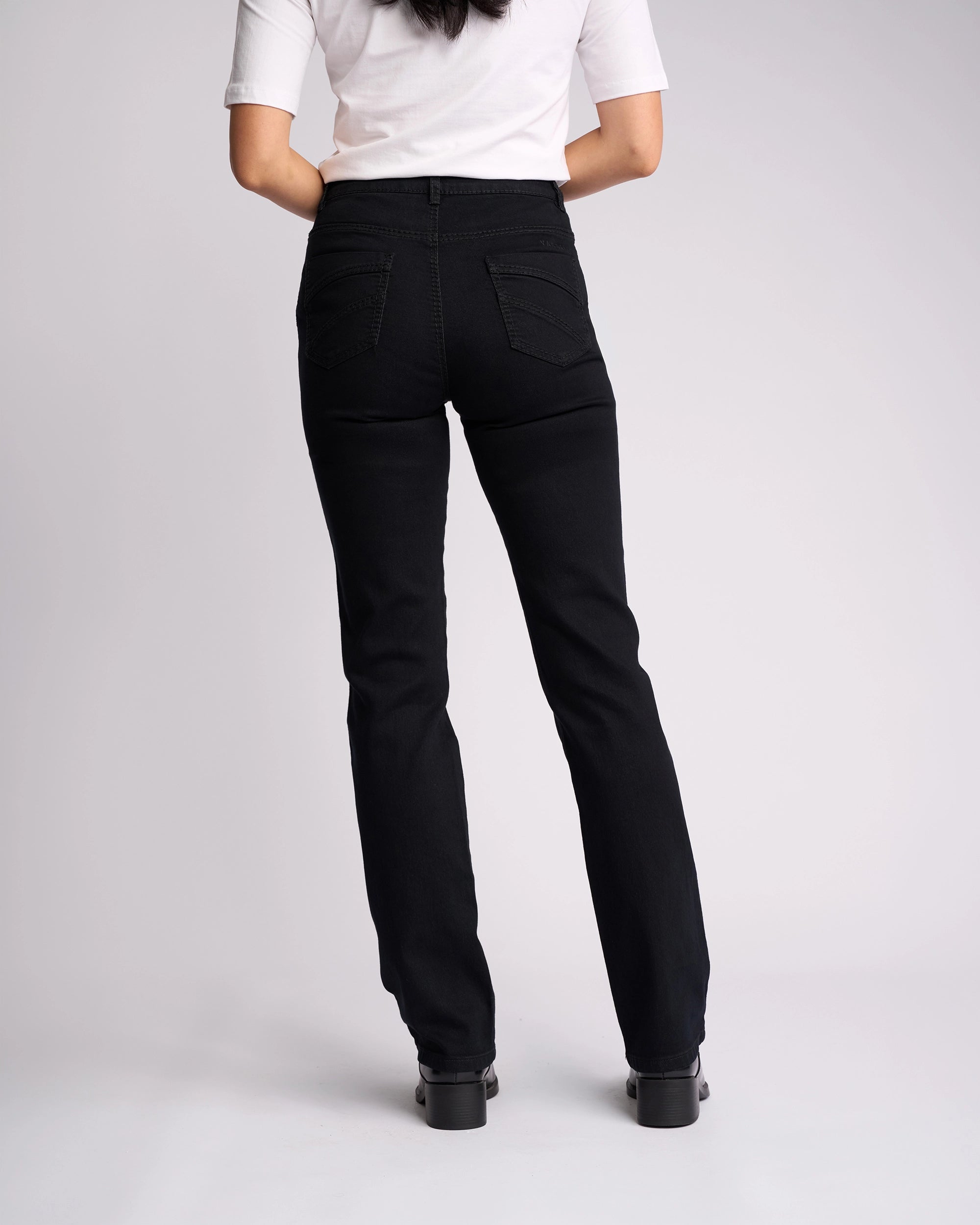 A woman faces away in a white short-sleeve top and Vassalli Tour Jean - Black Denim, featuring a high-rise waist and back pockets, paired with black shoes against a plain light background.