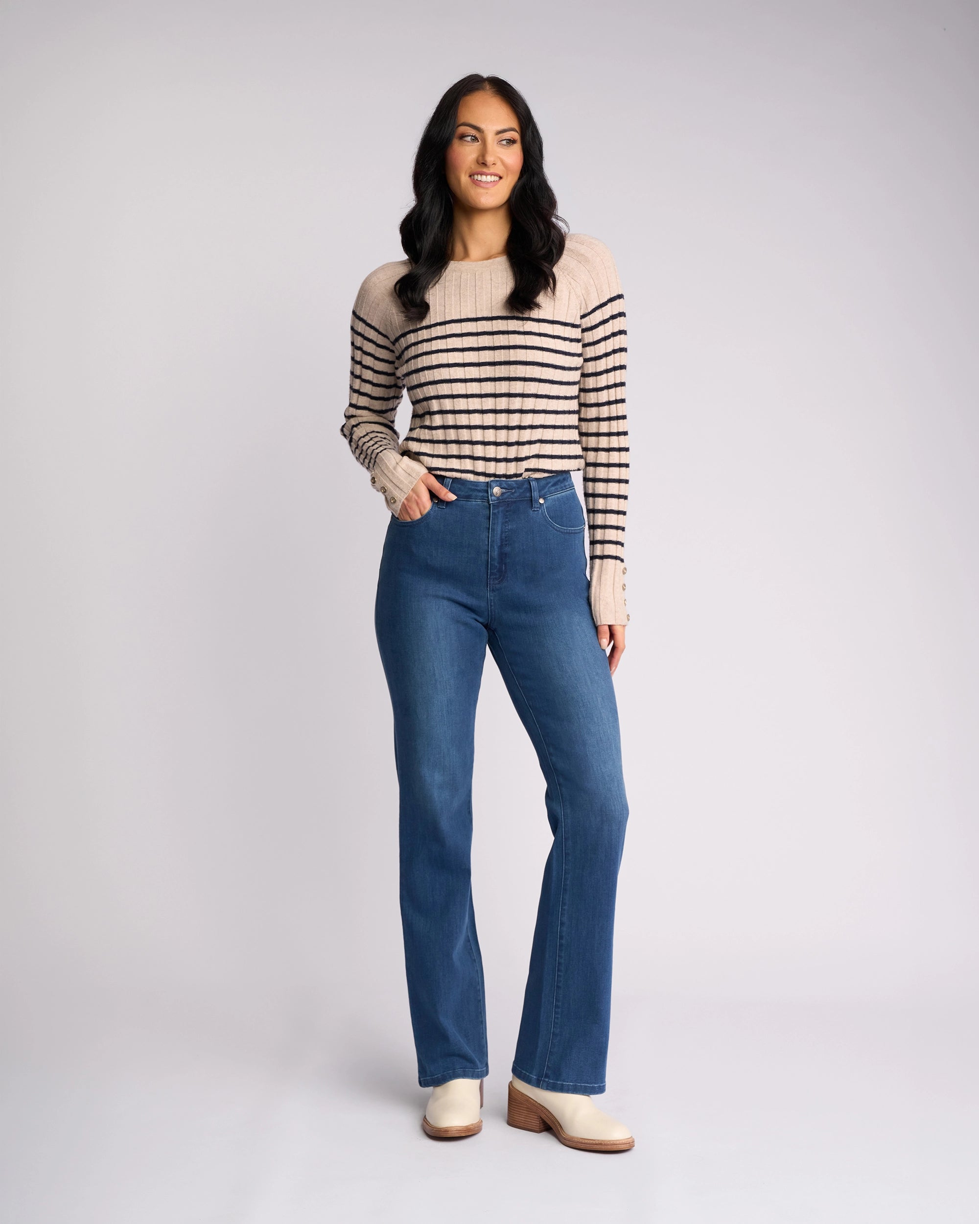 A woman with long dark hair smiles as she poses against a light background, wearing a beige sweater with black stripes, Vassalli Theory Jean - Blue Denim (With Fade), and off-white shoes.
