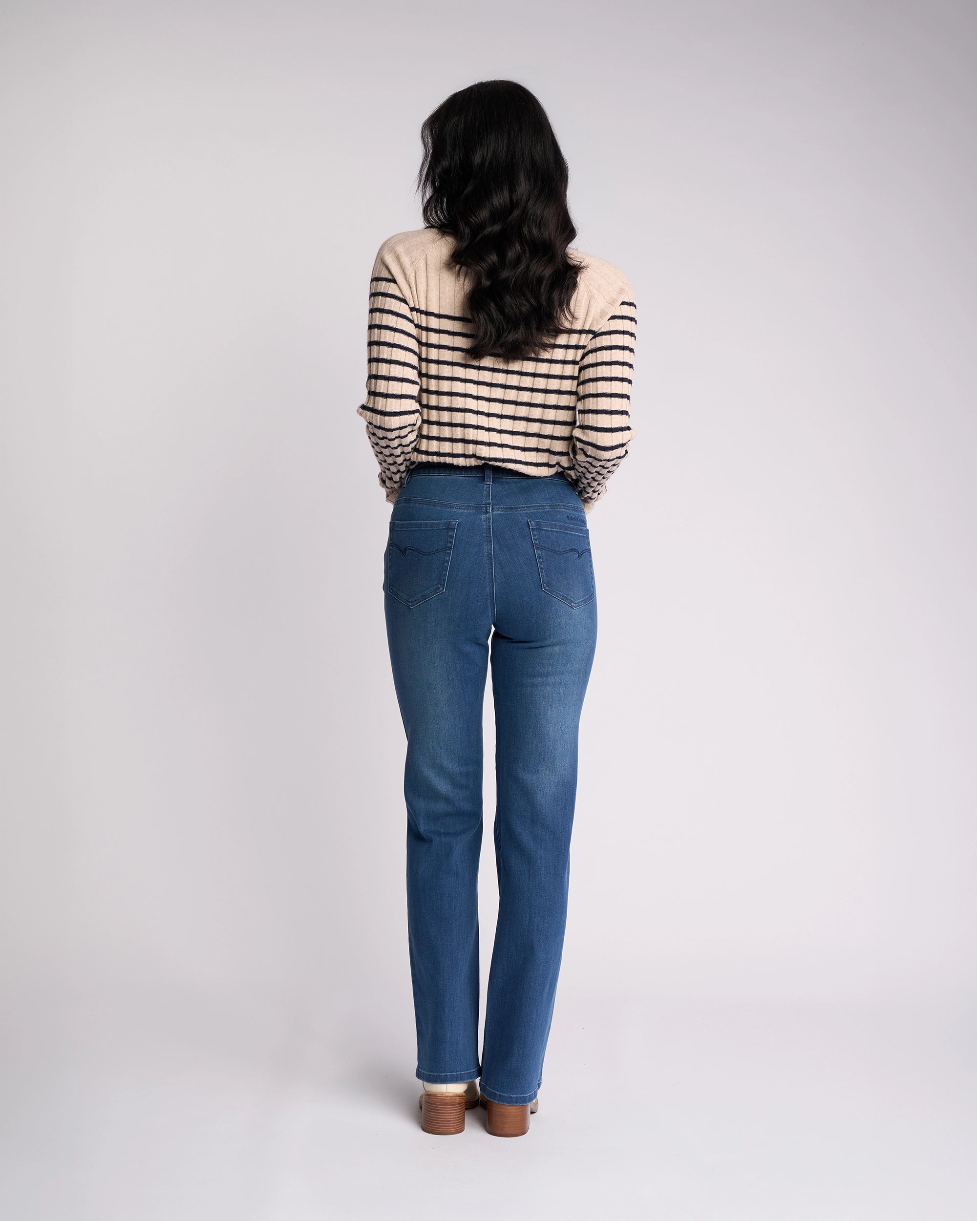 A woman with long dark wavy hair, facing away, wears a beige and black striped sweater and Vassalli's Theory Jean - Blue Denim (With Fade) against a plain light background, paired with brown heeled shoes.