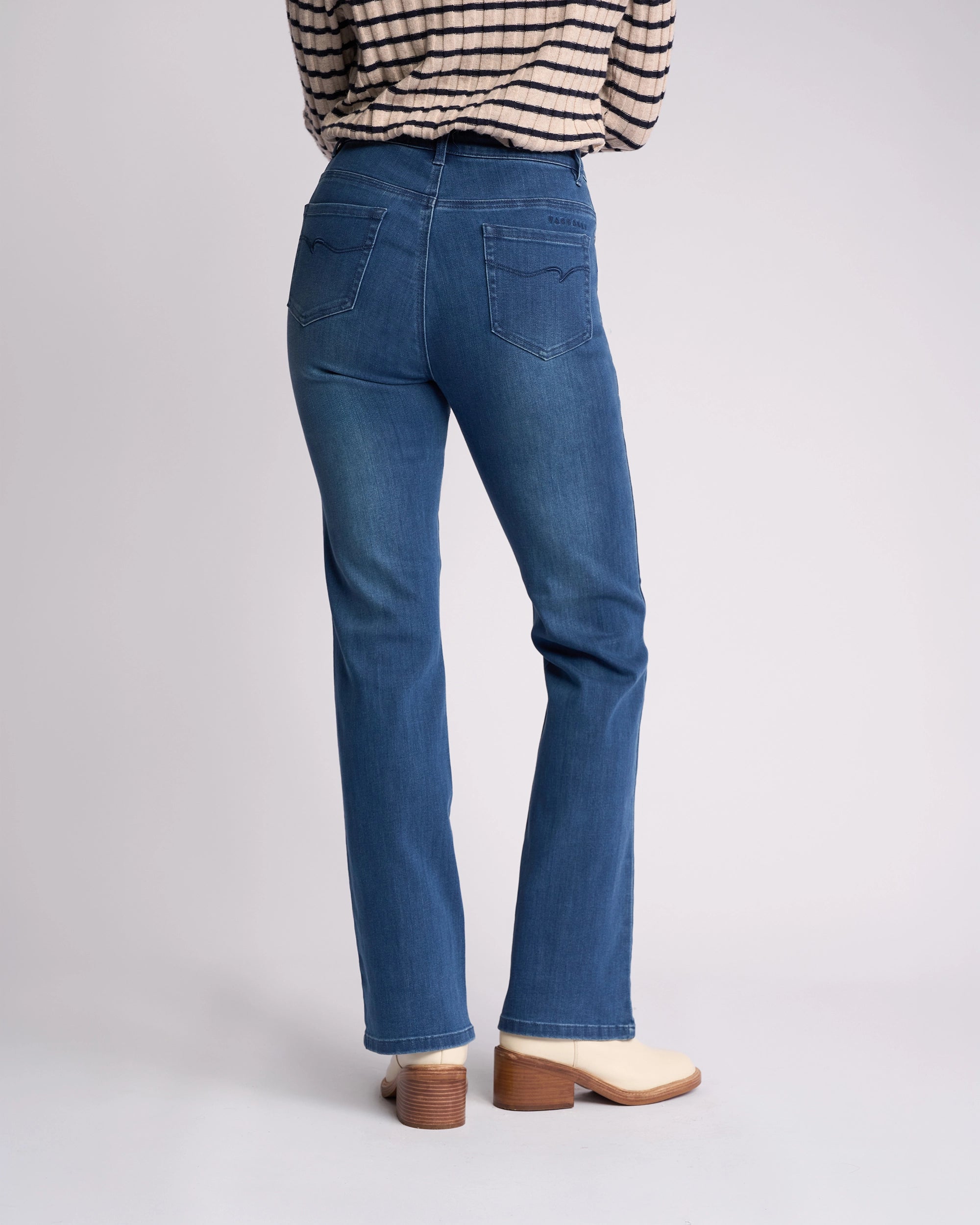 A person wearing Vassalli’s Theory Jean - Blue Denim (With Fade), a beige and black striped long-sleeve shirt, and cream heeled boots is shown from the back against a plain light background.